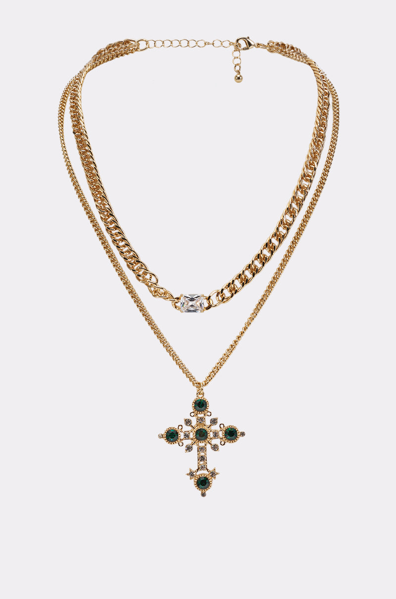The EMERALD SAINT CROSS LAYERED NECKLACE features both chunky and fine gold chains, showcasing a large ornate cross pendant embellished with green gemstones reminiscent of emerald rhinestones and accented with small pearls.