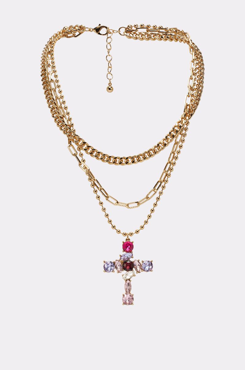 The JULIA NECKLACE features gold layered chains and a gold cross pendant adorned with red, purple, and clear gemstones, shown on a plain white background. 0