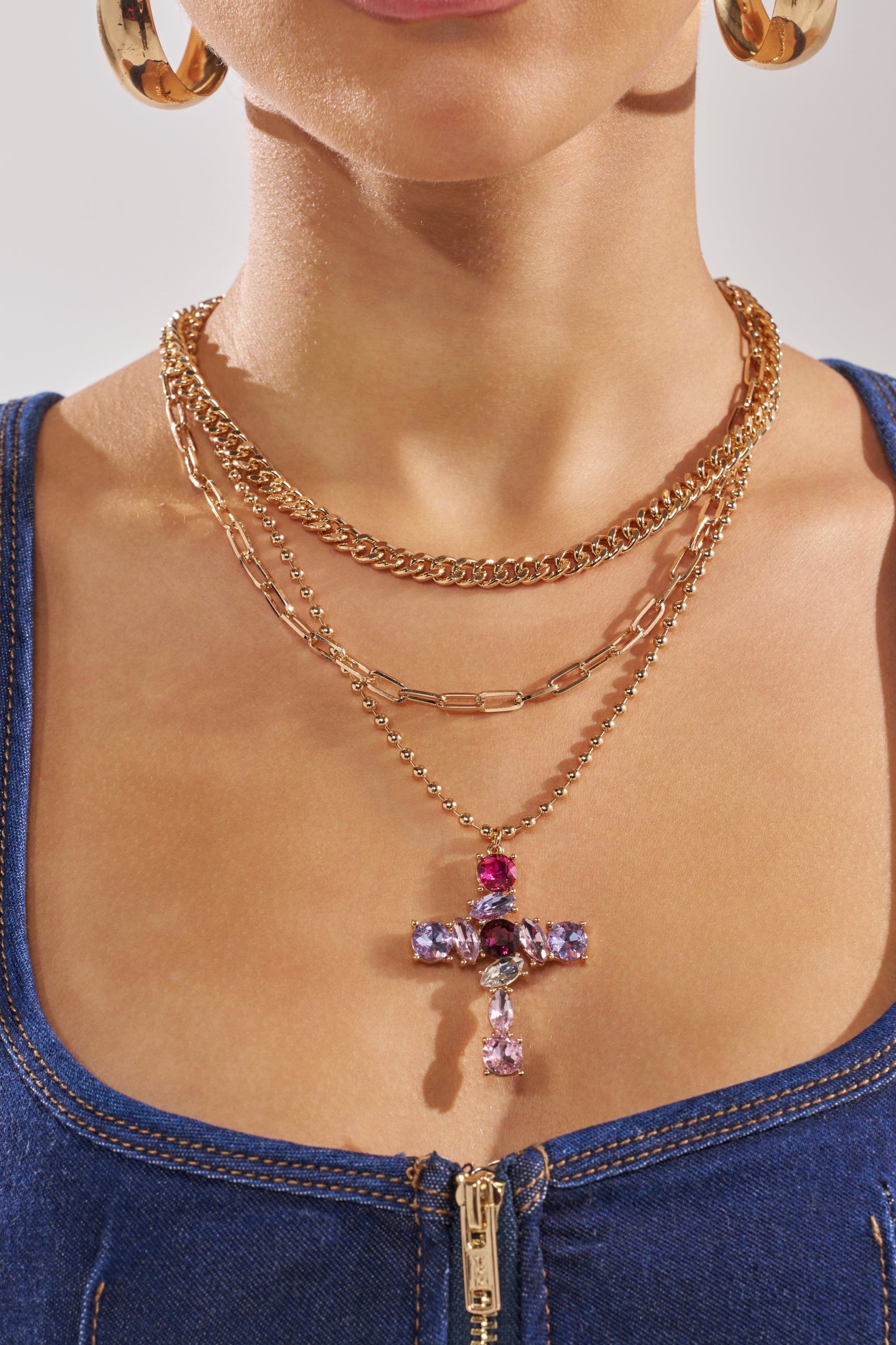 A woman wears the JULIA NECKLACE, a layered gold chain with a crystal cross charm, along with gold hoop earrings and a blue denim zip-up top. 1