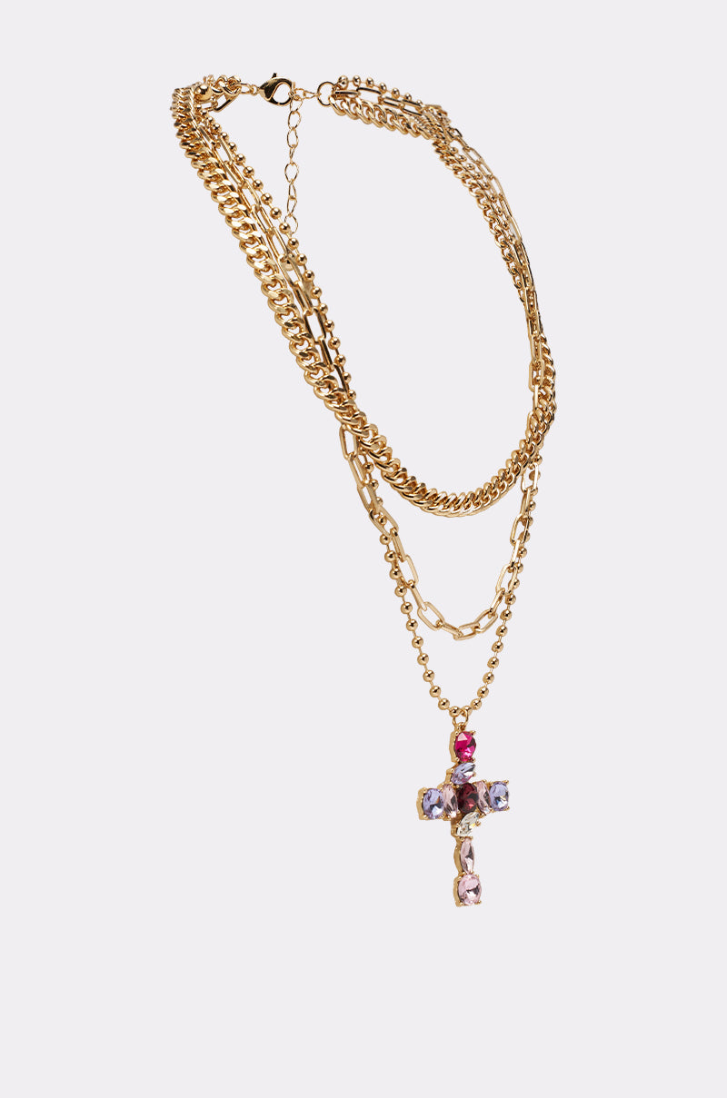 The JULIA NECKLACE is a gold layered chain featuring a crystal cross pendant with pink, purple, and clear gemstones, shown on a plain white background. 2