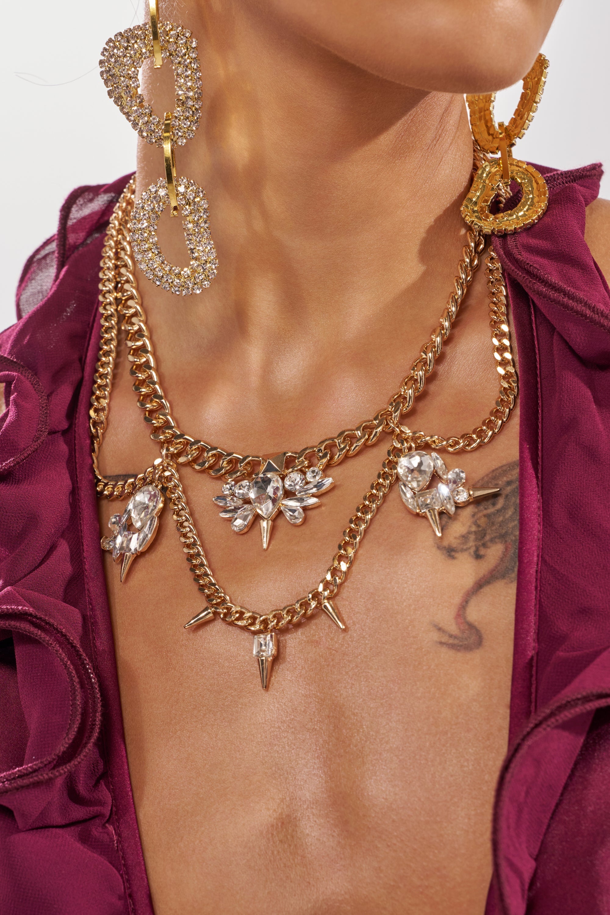 Someone wearing the SPIKEY NECKLACE, featuring multi-chain crystal cluster accents, paired with large sparkly gold hoops, a deep burgundy ruffled top, and a visible chest tattoo. 1