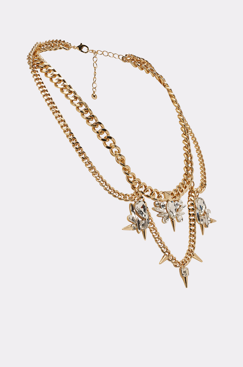 The SPIKEY NECKLACE showcases layered curb-link chains with chunky and delicate links, plus five crystal-studded pendants featuring gold spikes, all set against a plain light background. 2
