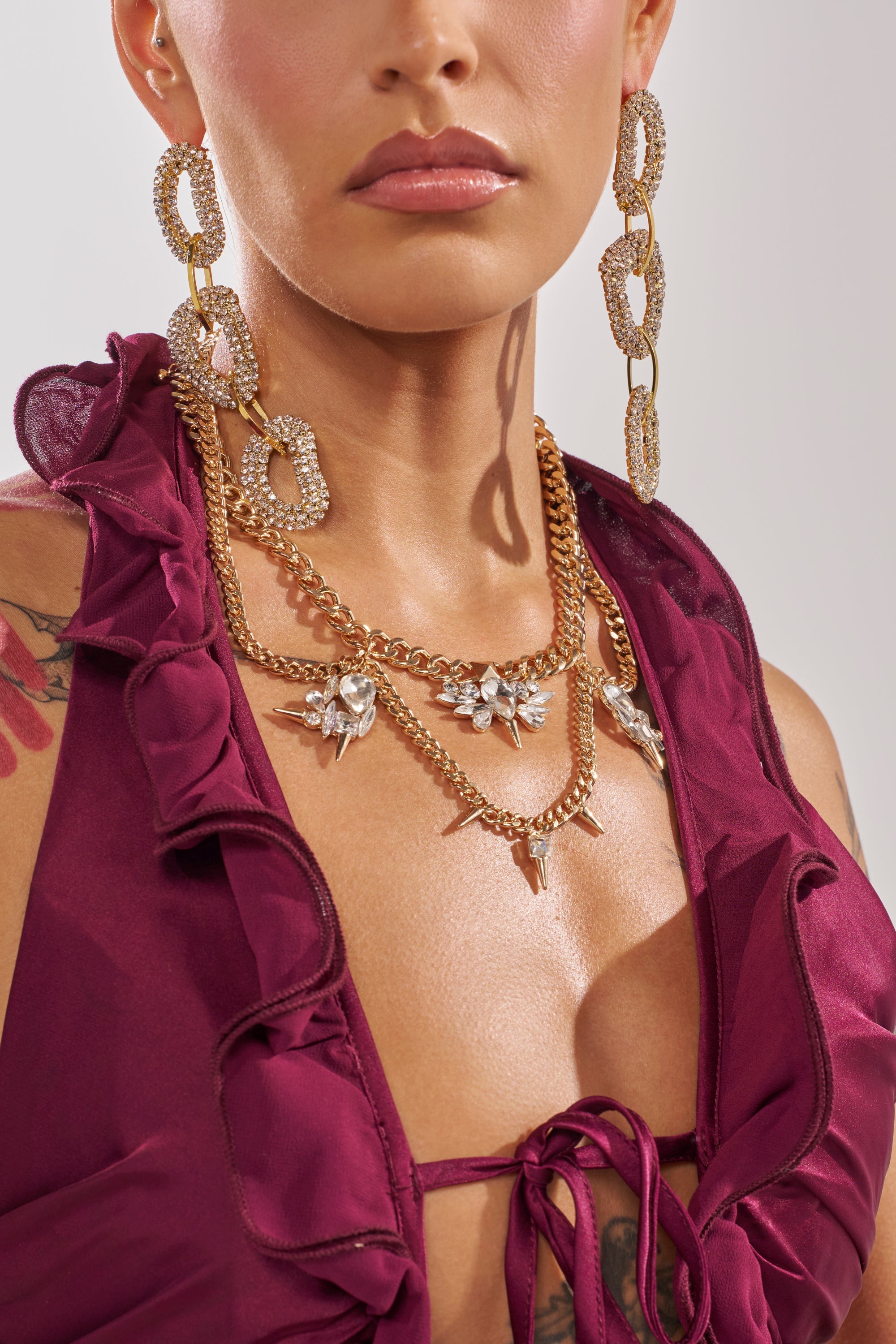 A woman wears the SPIKEY NECKLACE, a multi-chain statement piece with decorative pendants, paired with large gold and rhinestone earrings. She is dressed in a burgundy ruffled top with a front tie and has tattoos on her shoulder and chest. 4