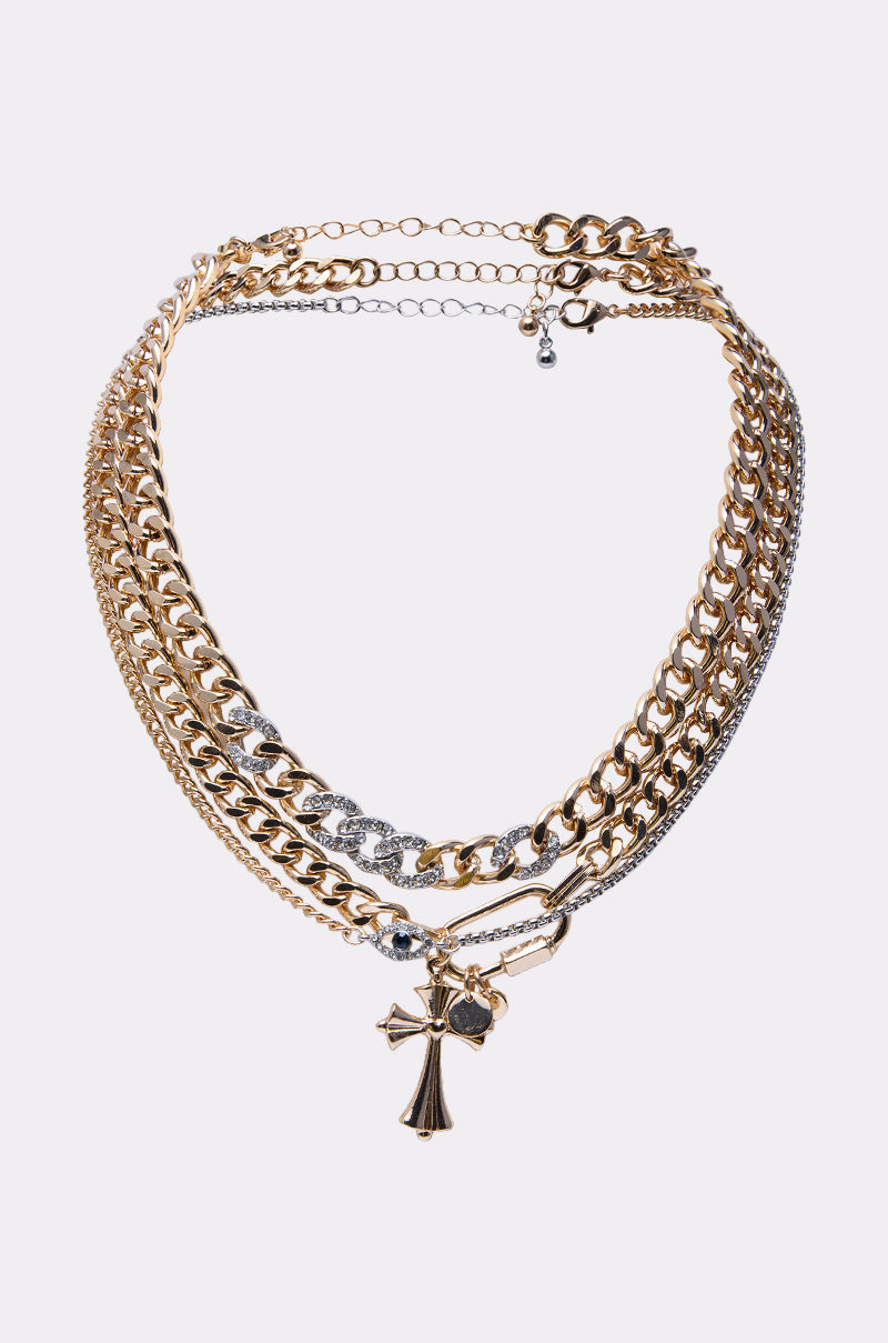 The HERE FOR IT NECKLACE SET features layered gold and silver chains with a bold cross pendant, combining chunky and delicate styles, all displayed against a plain white background. 0