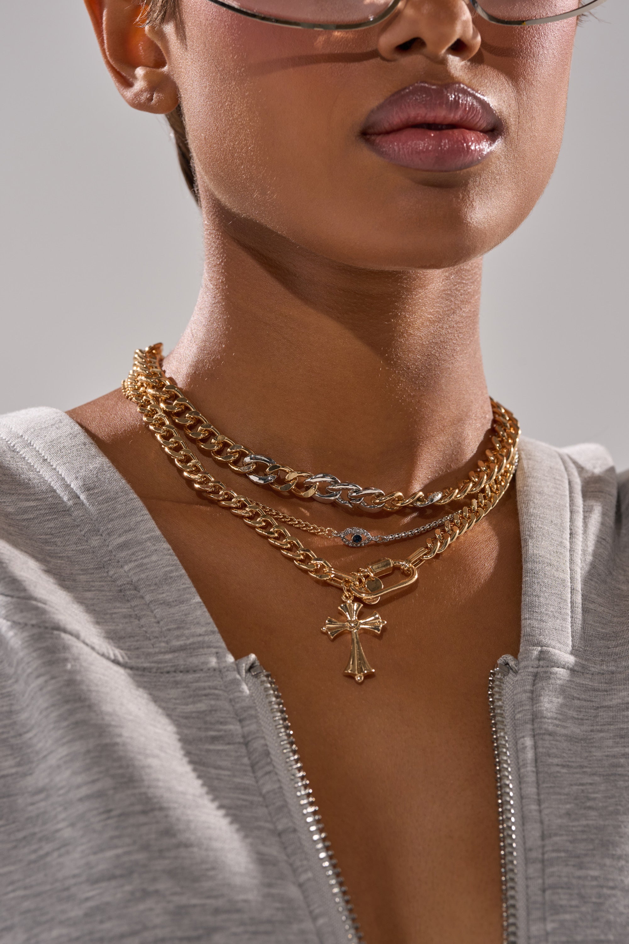 Wearing the HERE FOR IT NECKLACE SET—layered with a cross pendant and mixed metals—a woman in a grey zip-up top, glasses, and glossy lips is shown in a cropped image focusing on her lower face and neck. 1