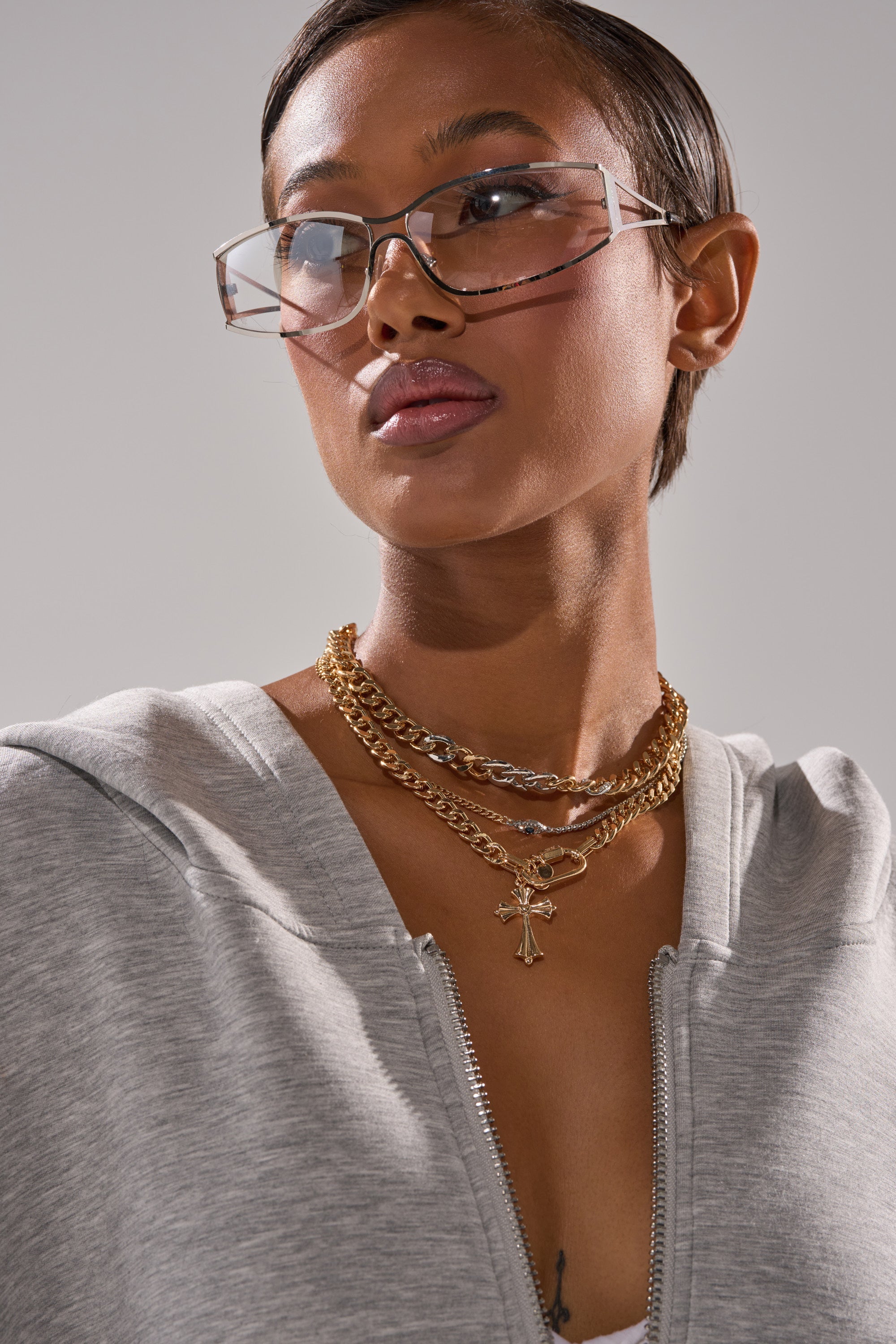A woman wearing angular clear glasses and the HERE FOR IT NECKLACE SET in gold tones, paired with a light gray zip-up top, looks to the side against a plain background. 2