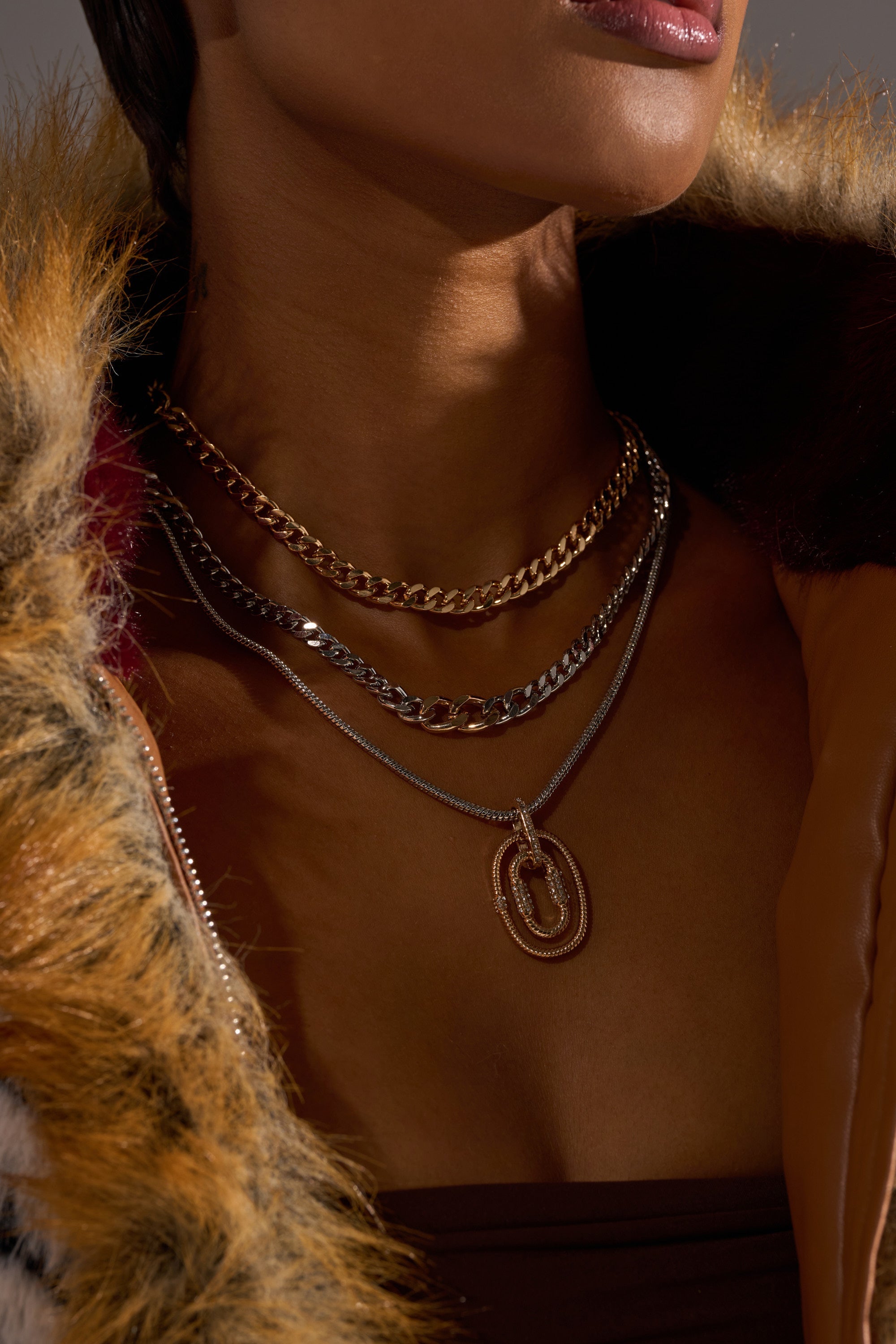 A close-up of a person wearing the CAN YOU NOT NECKLACE SET, featuring mixed metal chains with overlapping oval pendants, paired with a brown top and a faux fur-trimmed coat. The lower face and neck are visible. 1