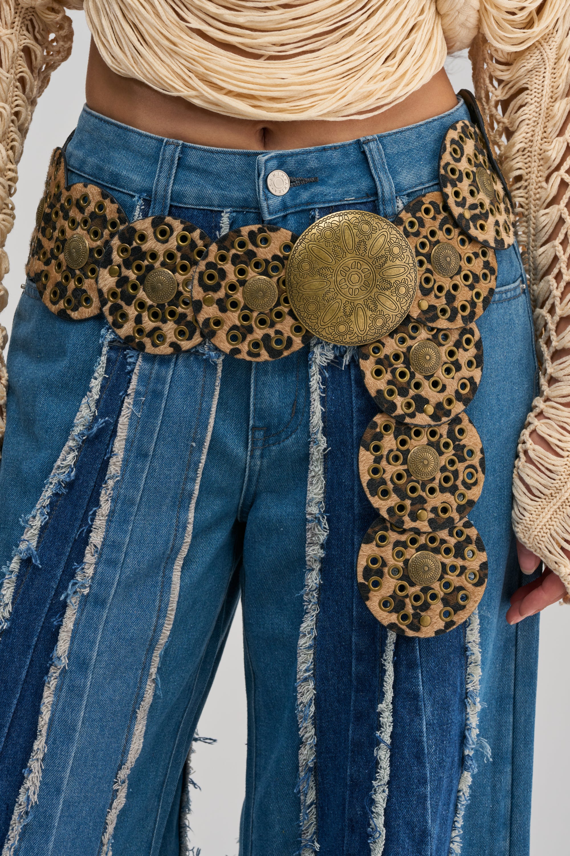 Person wearing high-waisted blue denim jeans with frayed stripes and the CHEETAH GIRL BELT—a wide, statement festival belt featuring ornate brass with circular floral designs—paired with a beige knit top with long sleeves.