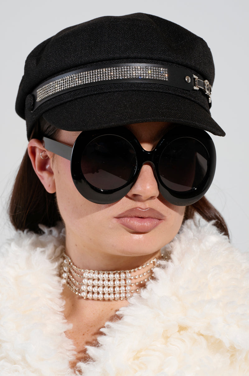 A person wearing oversized round sunglasses, an ITS GIVING BLING BAKER HAT adorned with rhinestone embellishments, and a fluffy white jacket. They have a pearl choker and a neutral expression against a plain background. 1