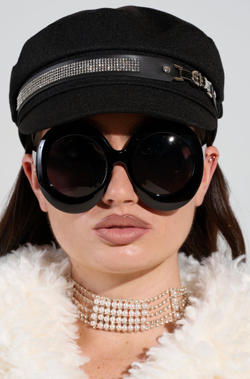 A person dons large round black sunglasses and a stylish accessory, the "ITS GIVING BLING BAKER HAT," featuring a rhinestone band. They are dressed in a fluffy white outfit, wear a layered pearl choker necklace, and have long dark hair set against a plain light-colored background. 2