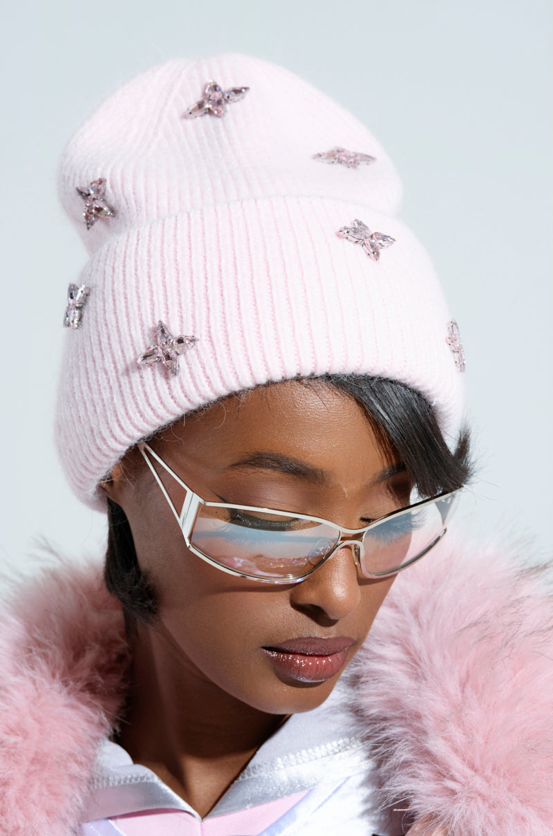 A person wearing a pink STAR POWER RHINESTONE BEANIE adorned with sparkling rhinestones and trendy angular sunglasses. The fur-trimmed hood of a light-colored jacket is slightly visible against a soft, neutral background.