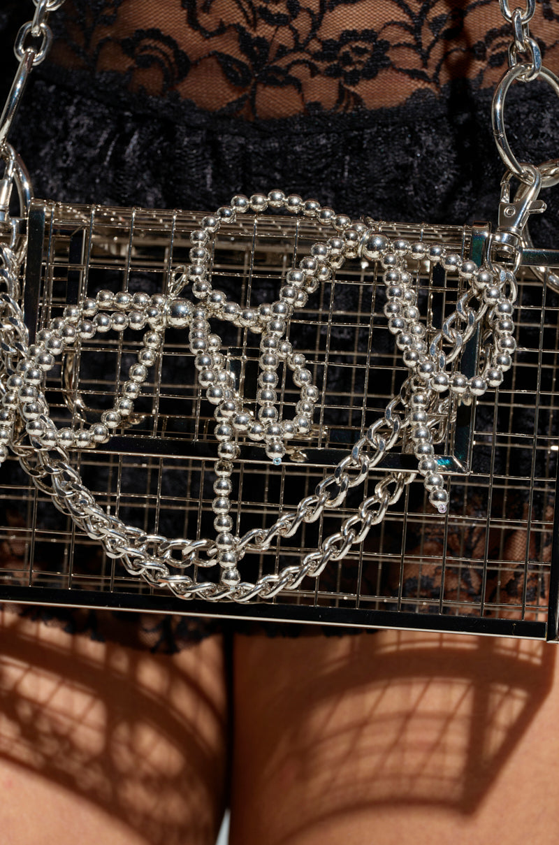 A close-up of a TRENDY CLIP ON CHARMS handbag, characterized by its rectangular metal design featuring a grid pattern and decorated with interwoven metallic silver beads and chains. The background displays a black lace garment.