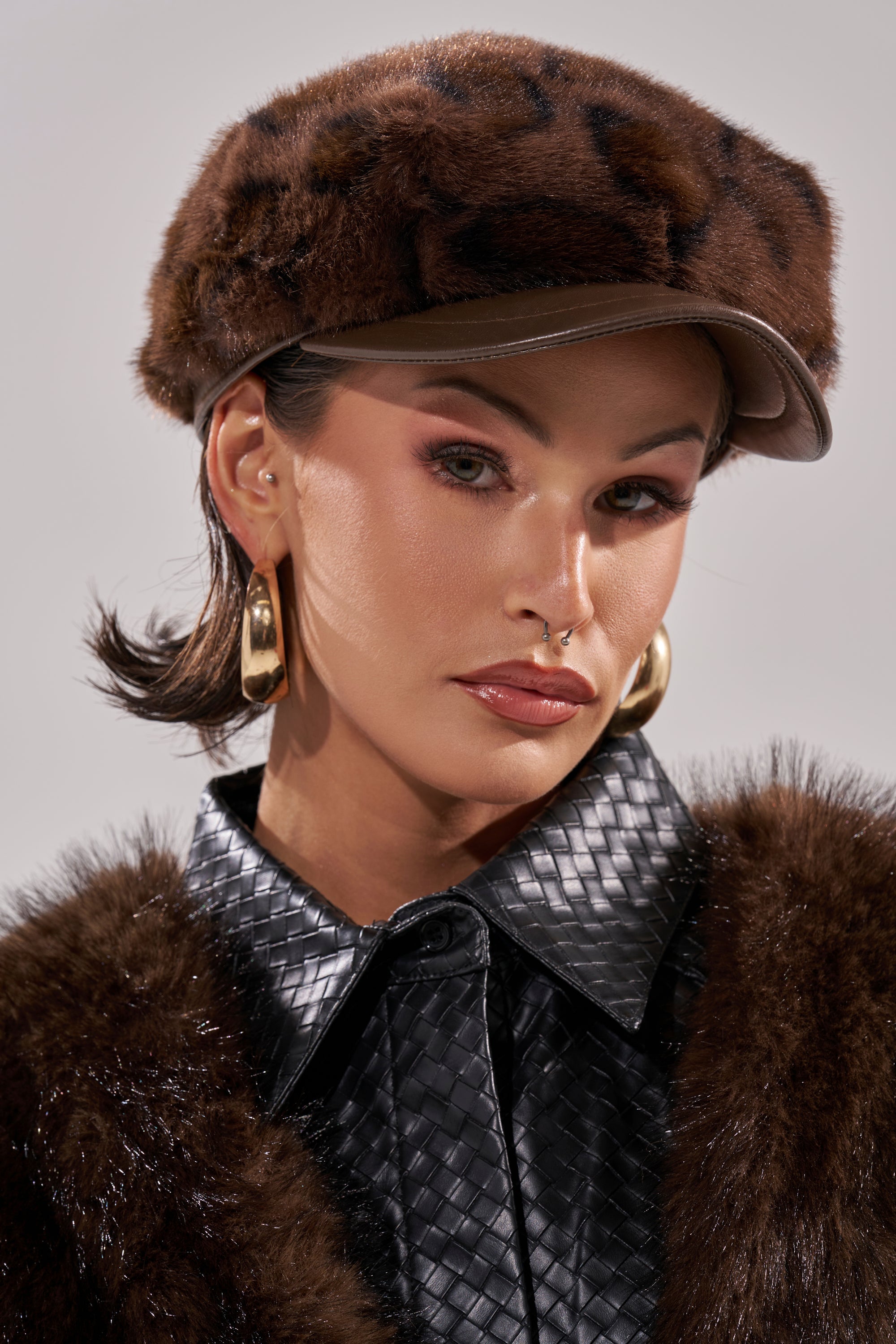 A person wears the I AM THE LUXURY FAUX FUR BRIM HAT, large gold hoop earrings, and a textured black jacket with a fur collar, gazing confidently at the camera against a neutral background. 0