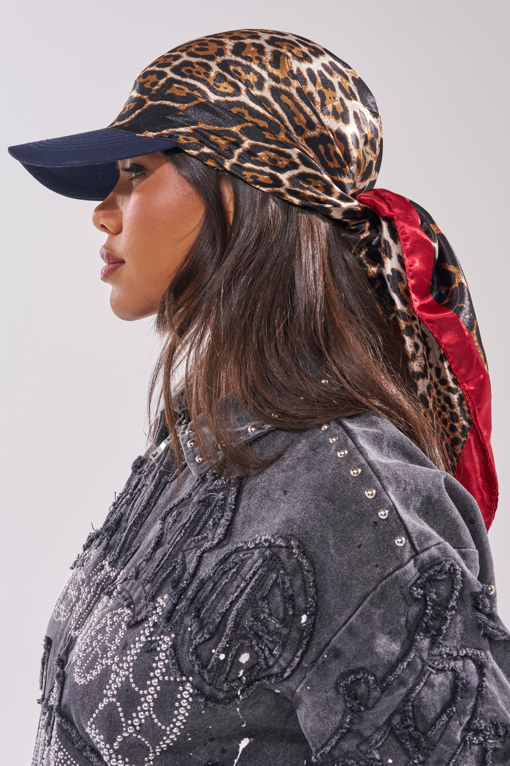 A woman in profile styles the WRAP ME AROUND HAT—a leopard print and red scarf over a black cap—with a sparkling black denim jacket, creating a bold travel look against a plain, light background.