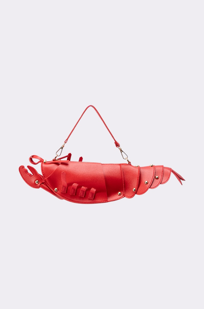 The EAT ME PURSE is a red faux leather handbag shaped like a lobster, accented with gold rivets and a matching red strap. Displayed on a plain light background, this crawfish-inspired purse is the perfect novelty summer accessory.