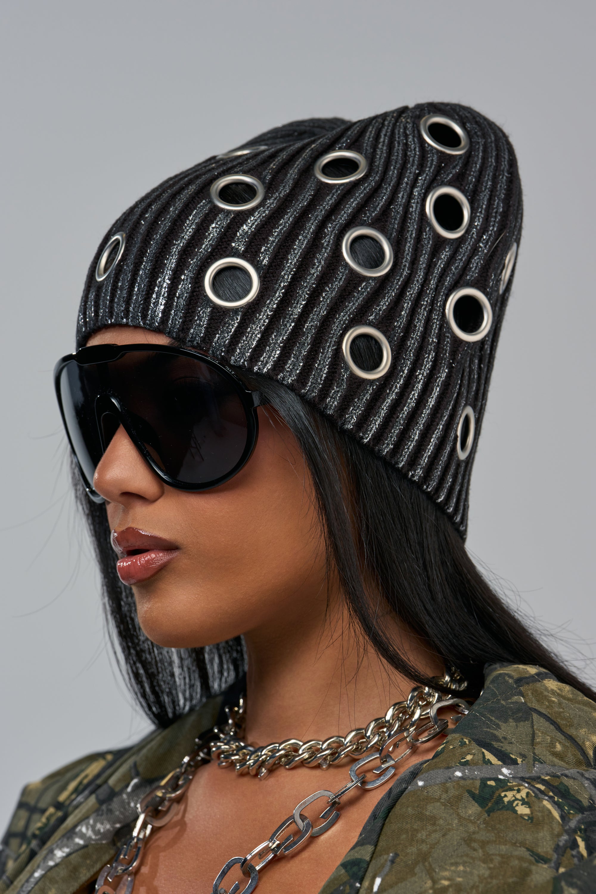 A person wearing a FINAL COUNTDOWN HAT, an oversized metallic knit beanie embellished with silver eyelets, and large wrap-around sunglasses.