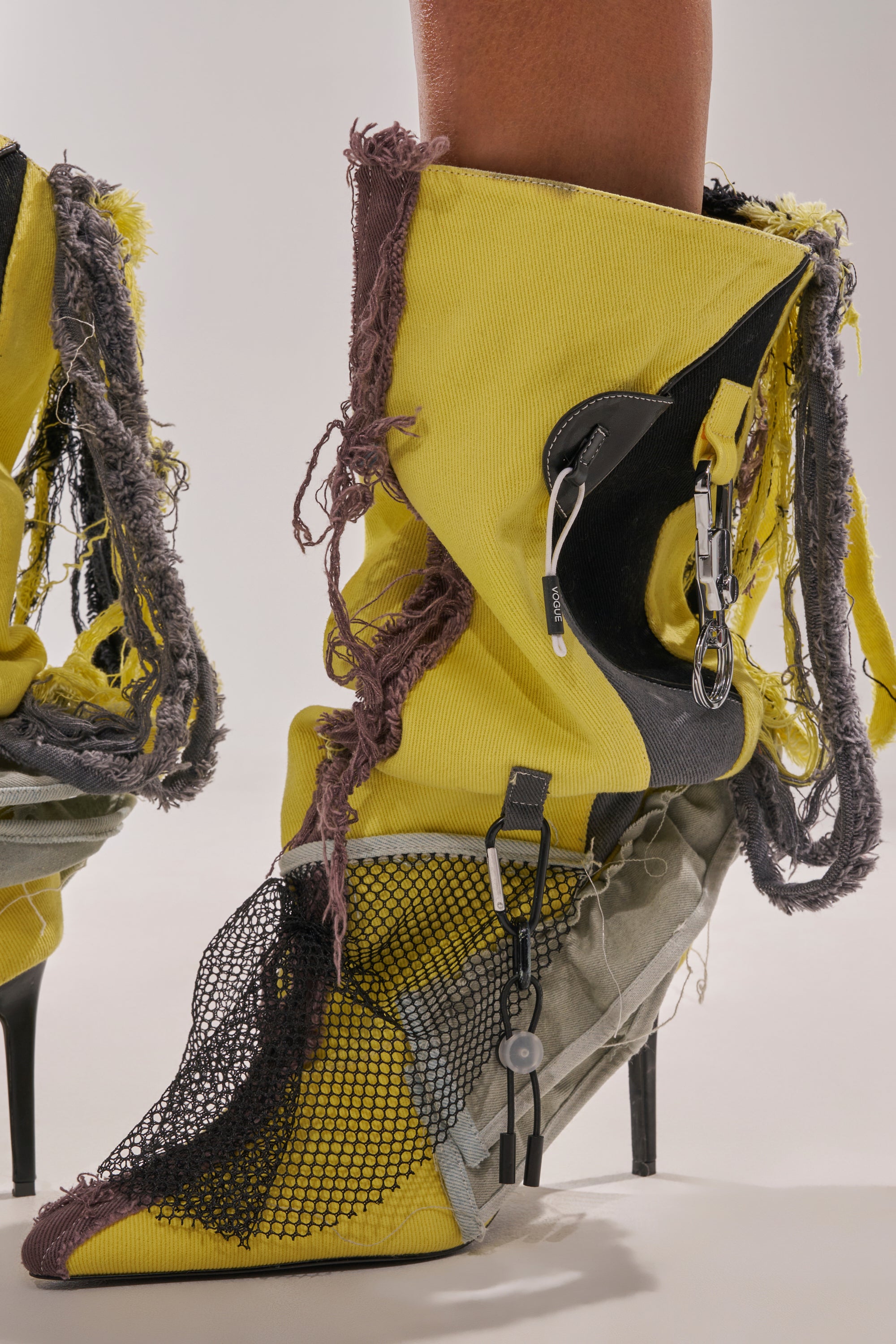 A close-up of a person wearing the AKIRA GOLD COALESCE SPORTY MIXED MATERIAL STILETTO BOOT: yellow knee-high boots with patchwork gray and black fabric, frayed edges, mesh details, zippers, and metallic hardware. 7