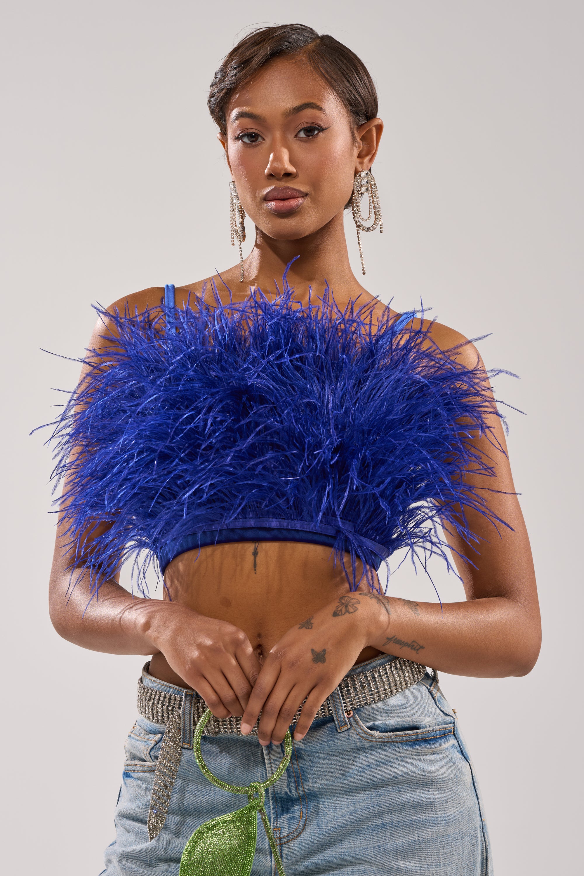 A woman poses confidently in the BRIGHT LIGHTS FEATHER TOP—royal blue, velvet, cropped—paired with light blue jeans, sparkling earrings, and a green glittery purse. Her short hair is styled perfectly, with tattoos visible on her hands. 0