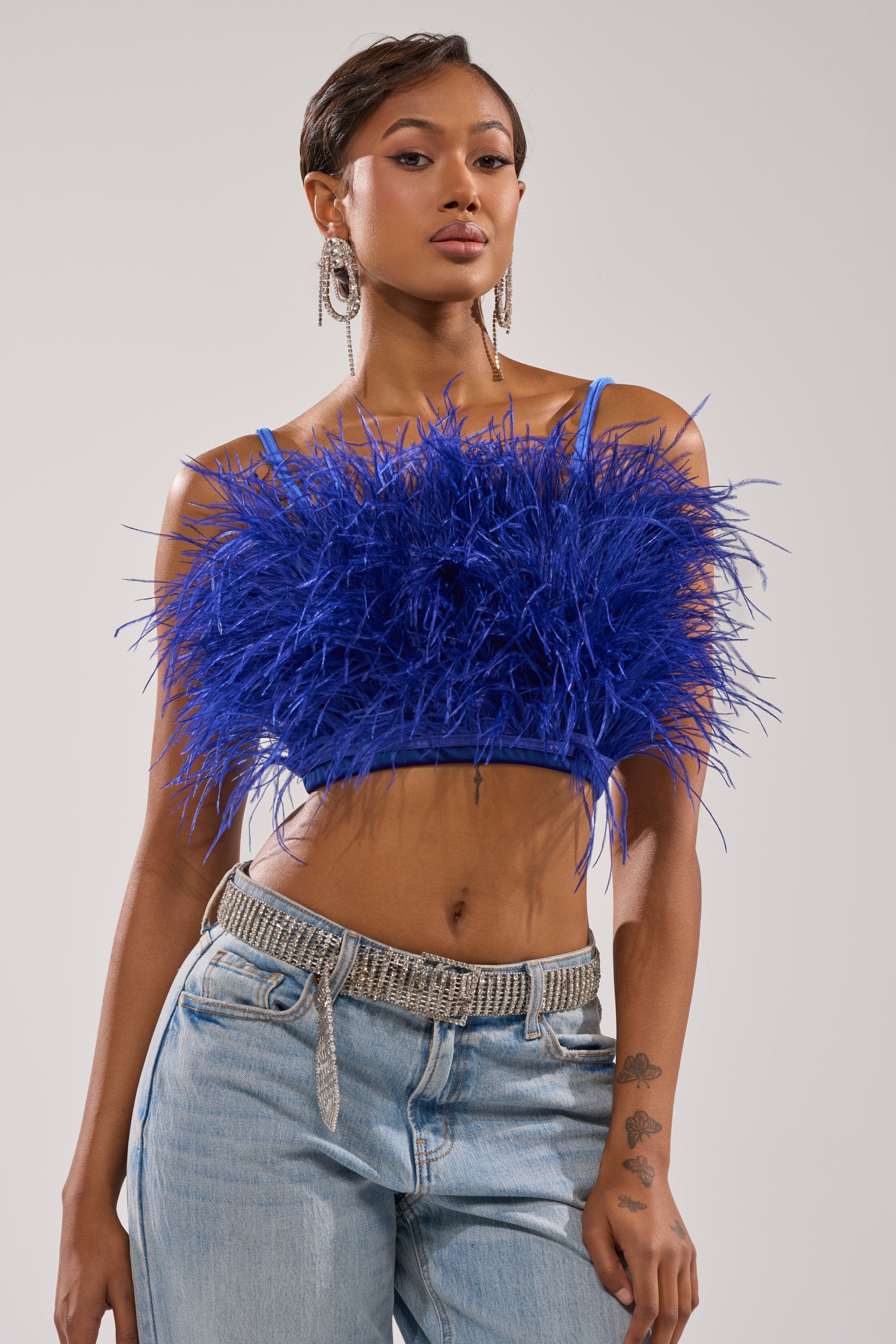 A woman with short hair poses confidently against a plain light background, wearing the BRIGHT LIGHTS FEATHER TOP in royal blue velvet, cropped fit, paired with light blue jeans, a silver chain belt, and dangling earrings. 2
