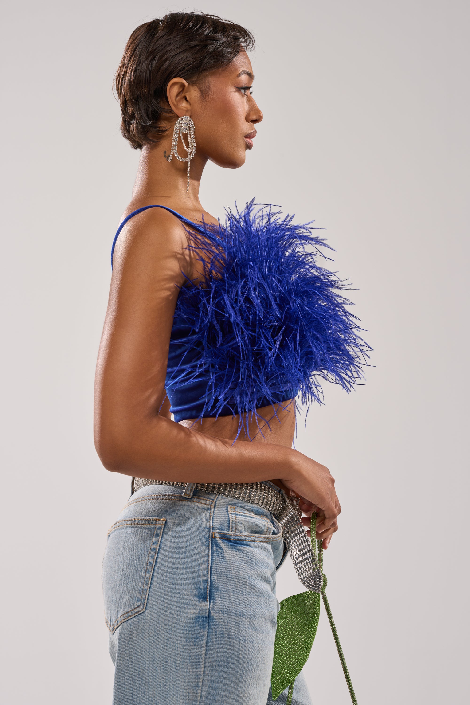 A woman in profile wears the BRIGHT LIGHTS FEATHER TOP in royal blue velvet with a cropped fit, paired with light blue jeans and bold earrings. She holds a green stem, her short hair standing out against a plain light background. 3