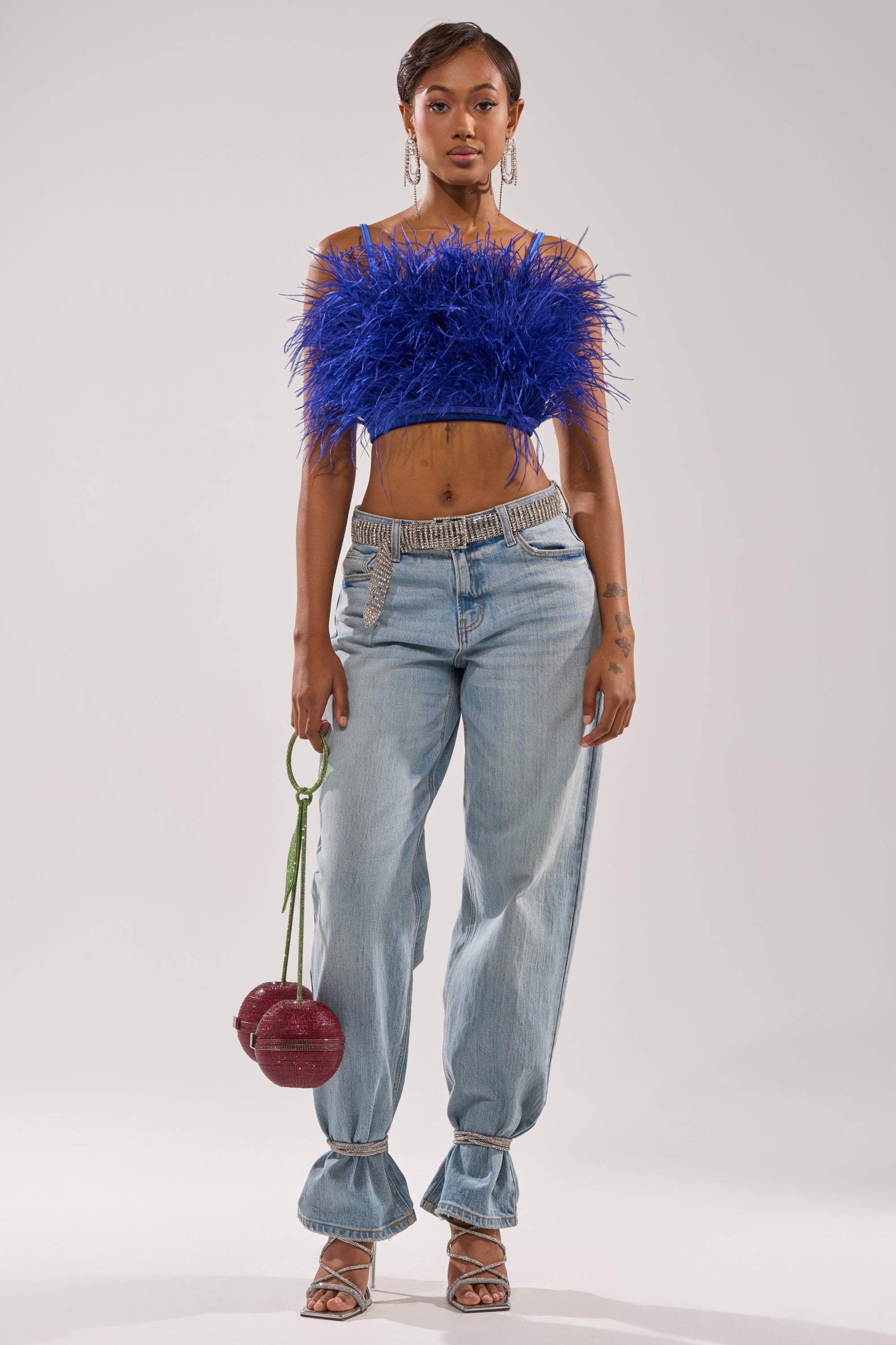 A woman with short hair and hoop earrings poses against a plain background wearing the BRIGHT LIGHTS FEATHER TOP in royal blue velvet, high-waisted light blue jeans, strappy heels, and carrying a red spherical purse. 5