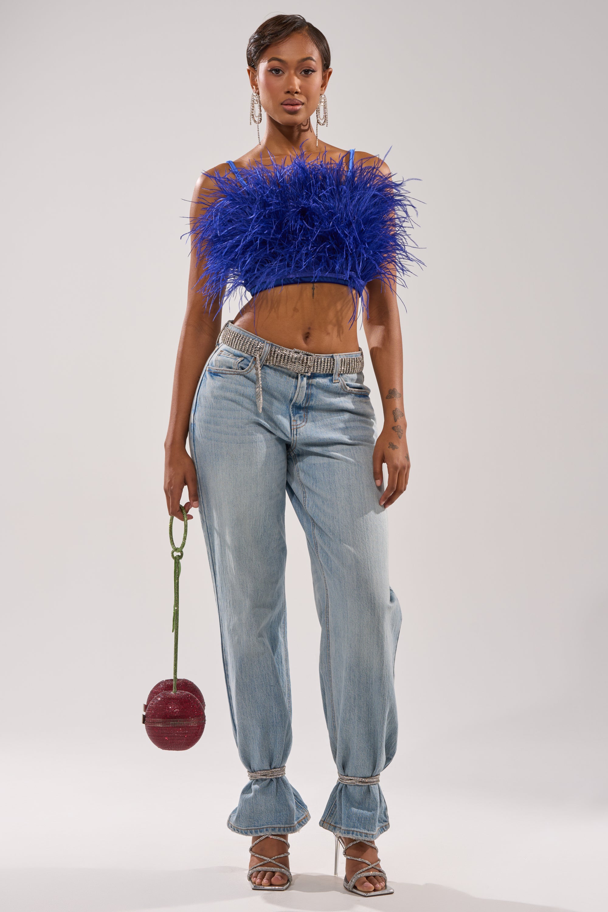 A woman faces forward in a royal blue BRIGHT LIGHTS FEATHER TOP, light blue loose-cuff jeans, strappy heels, and holds a round red handbag. The background is plain and light-colored. 6