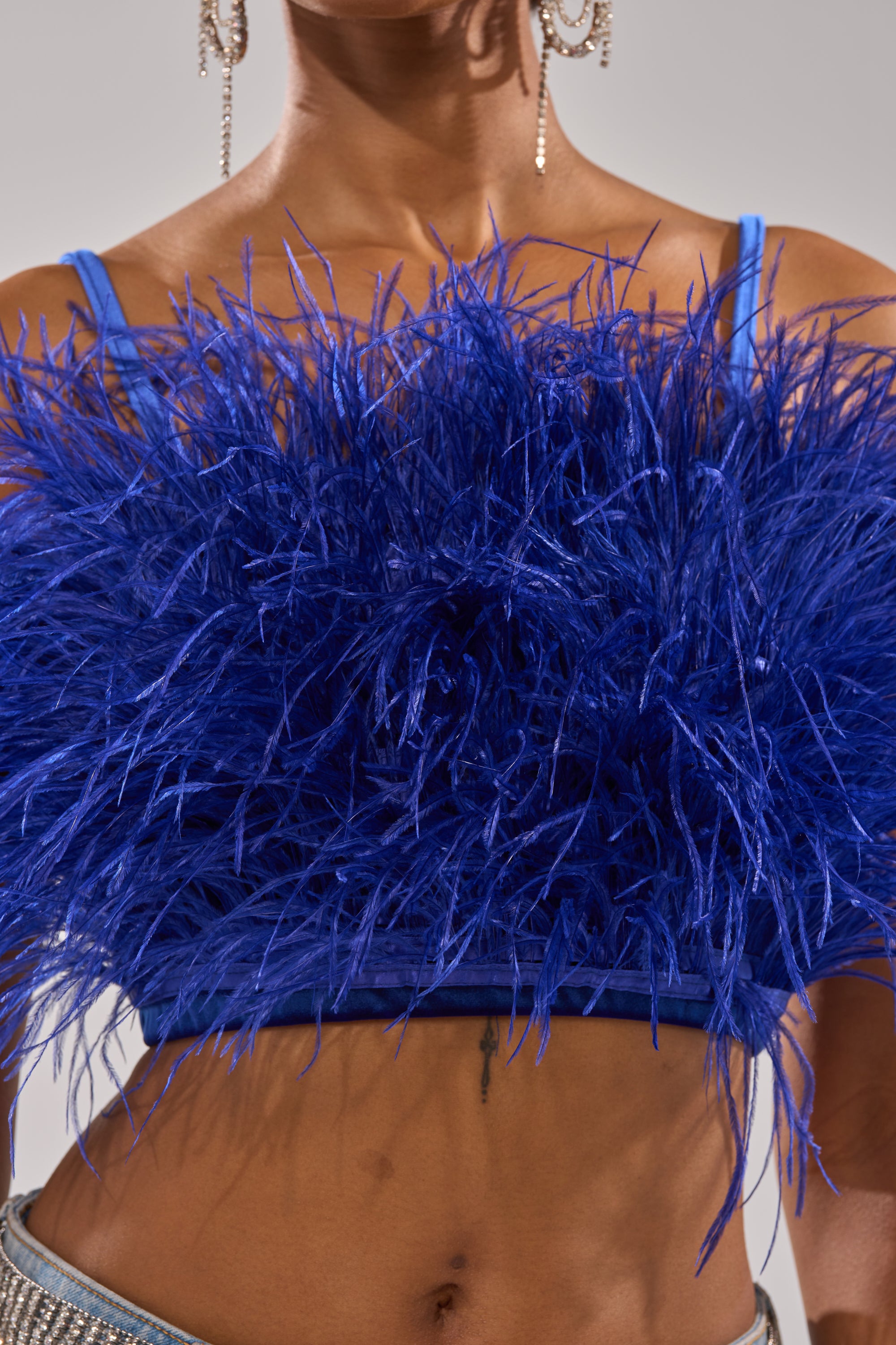 Wearing the BRIGHT LIGHTS FEATHER TOP in royal blue velvet with a cropped fit and dramatic fluffy feathers, only the lower half of their face and torso are visible. Elegant dangling earrings complete the look. 7