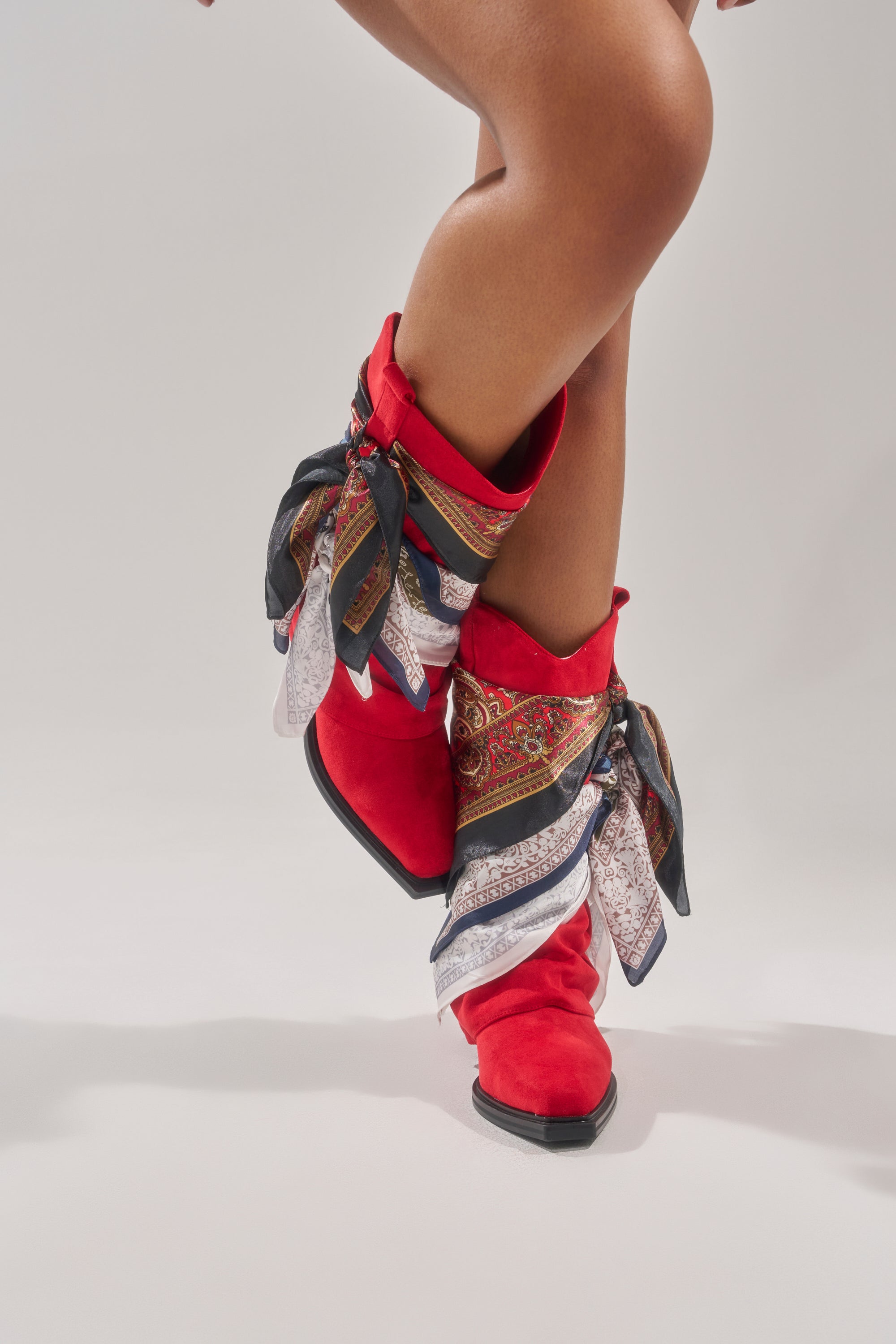 Wearing the AZALEA WANG COBALT RED FAUX SUEDE SCARF EMBELLISHED WESTERN BOOTIE, a person stands with legs crossed against a light gray background. The booties feature patterned silk scarves tied around the ankles. 0