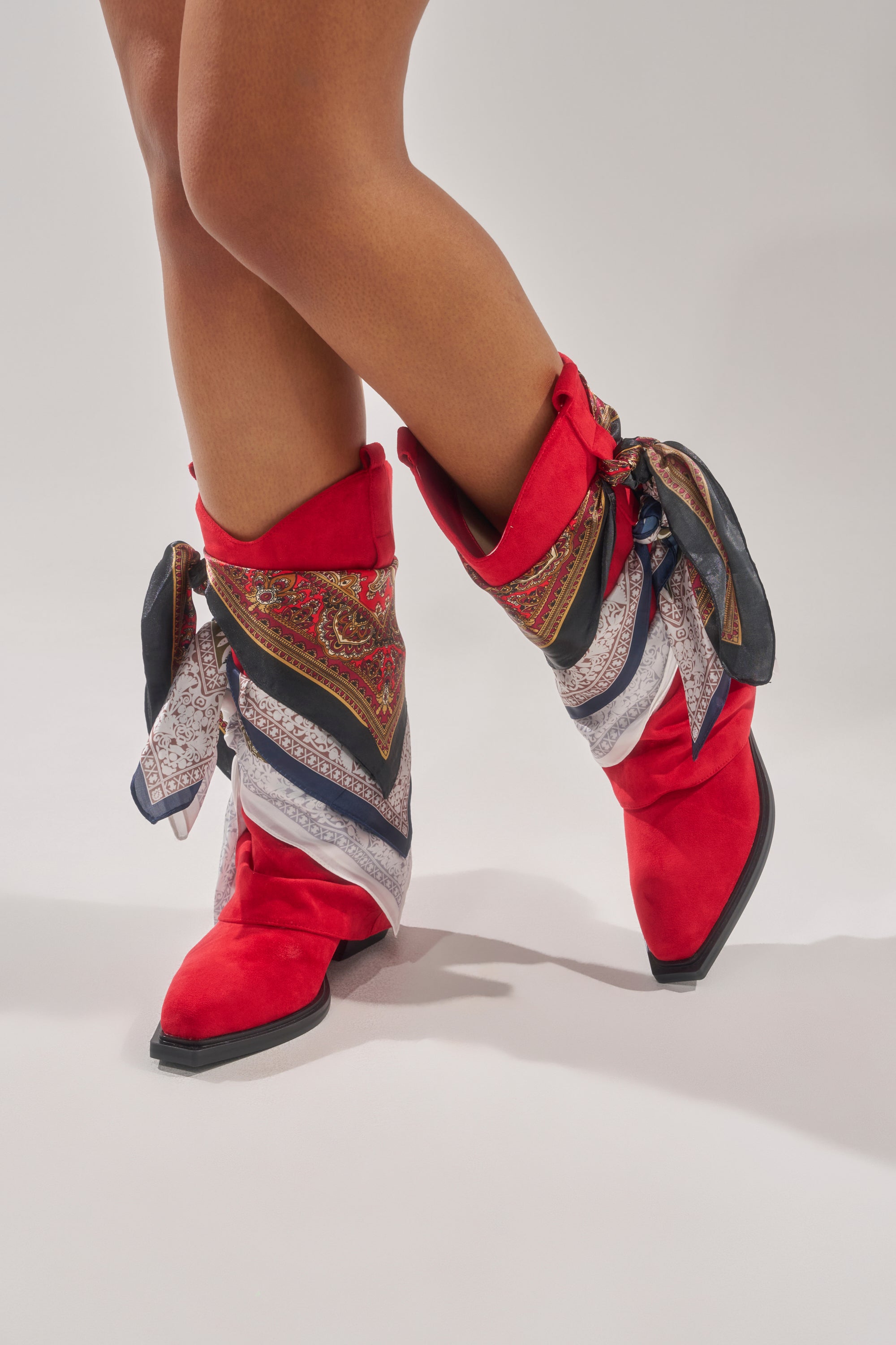 A person stands on a light gray surface wearing AZALEA WANG COBALT RED FAUX SUEDE SCARF EMBELLISHED WESTERN BOOTIES with pointed toes and colorful patterned scarves tied around the ankles. 1