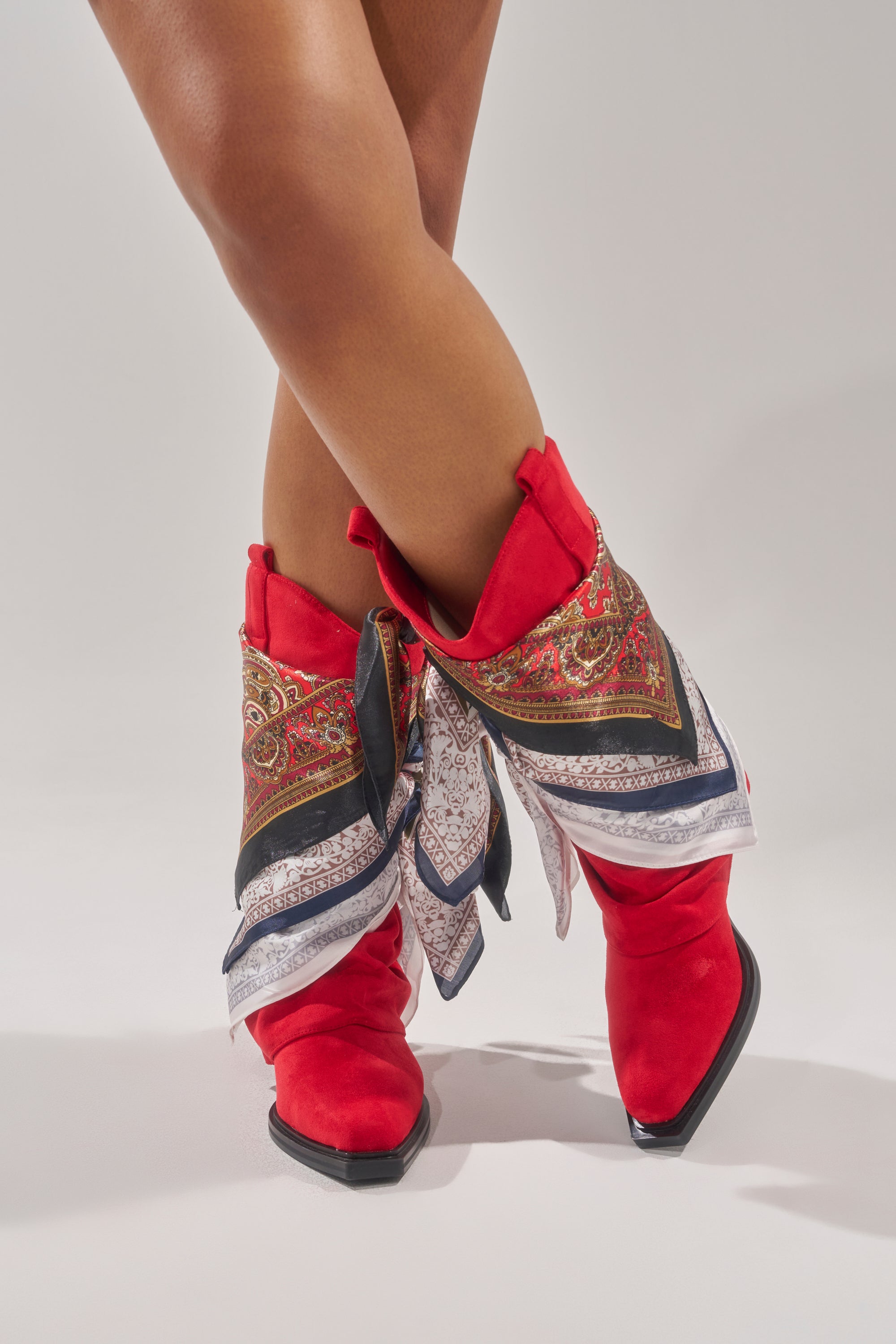 A person stands cross-legged against a light gray background, wearing AZALEA WANG COBALT RED FAUX SUEDE SCARF EMBELLISHED WESTERN BOOTIES with patterned silk scarves tied around each boot. 2
