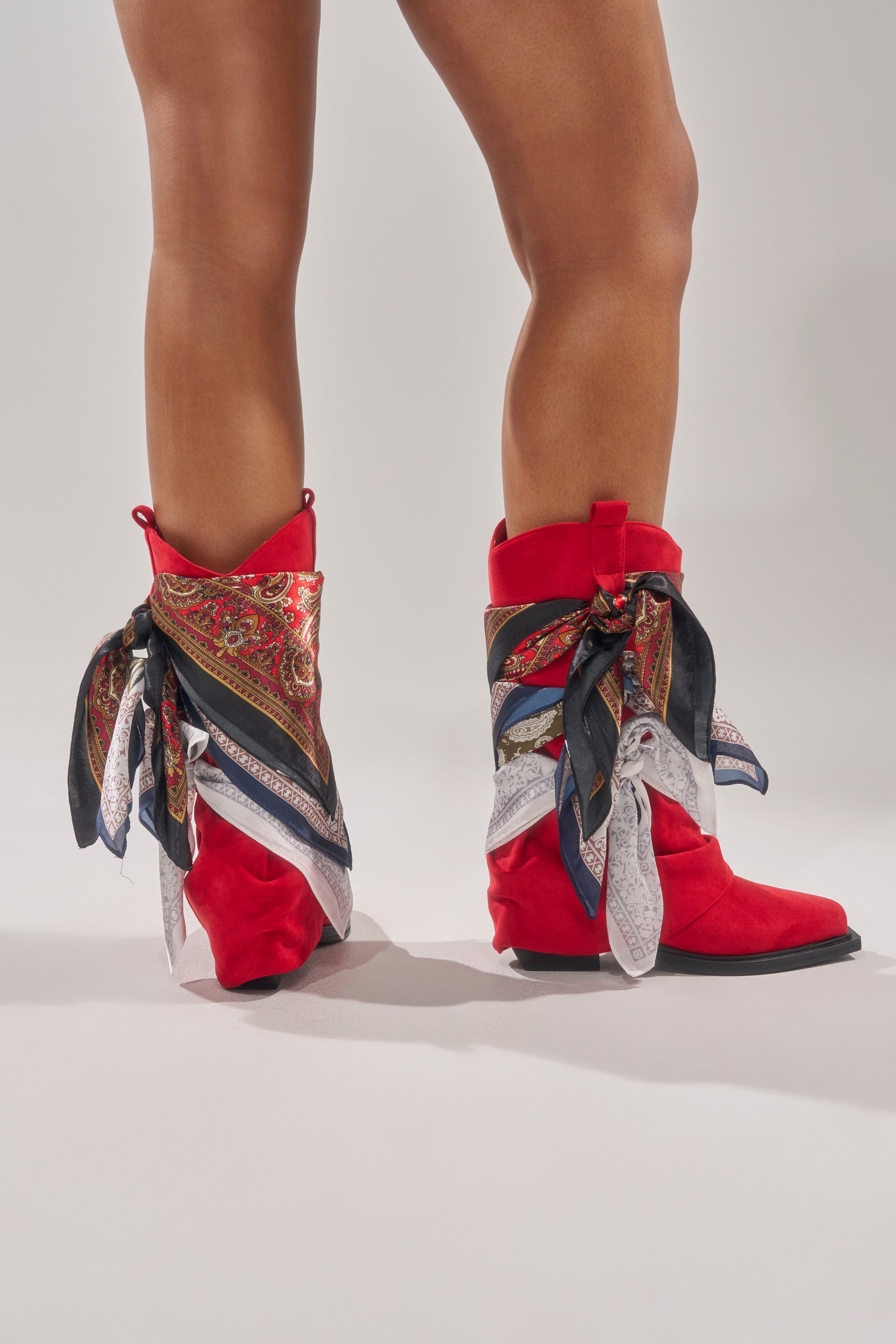 Someone stands on a light gray surface wearing the AZALEA WANG COBALT RED FAUX SUEDE SCARF EMBELLISHED WESTERN BOOTIE, featuring patterned silk scarves tied around the ankles. 3
