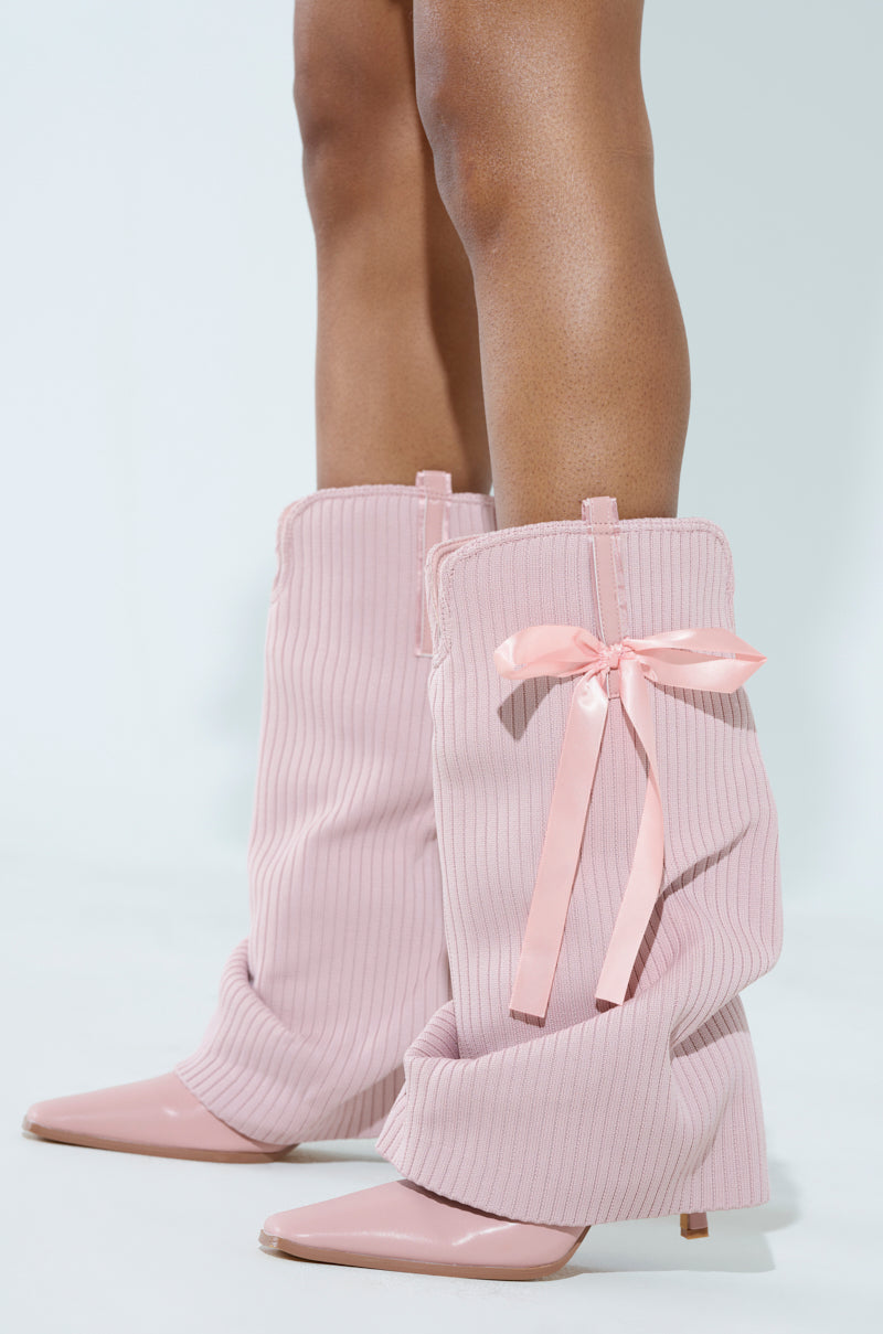 Sporting the CODEWORDS Pink Bow Stiletto Fold Over Boots, featuring oversized, ribbed fabric, pointed heels, and charming pink ribbon bows on the side, this stylish vegan leather footwear pops against a light pastel backdrop for a fashionable blend of elegance and flair.