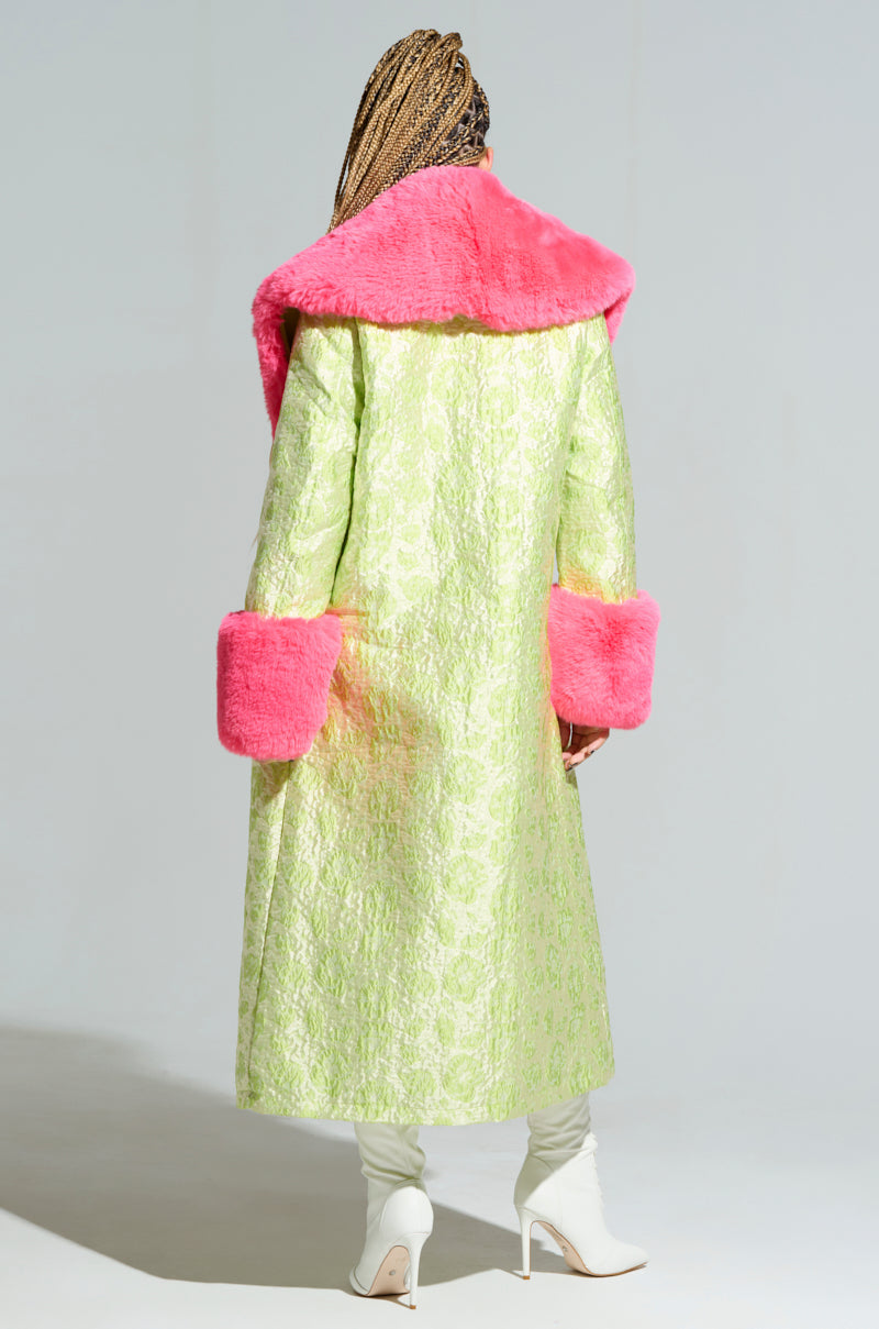 Facing away, a person with braided hair is wrapped in the luxurious Color Me Pretty Brocade Trench, distinguished by its bright pink faux fur collar and cuffs. Complementing this look are white heeled boots. The plain gray background enhances the vibrant Y2K-inspired accessories. 4