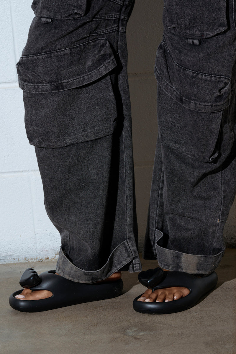 A person is wearing dark gray cargo pants with large pockets, paired with AKIRA COMPASSION BLACK SANDALS featuring an EVA upper. The cuffed pants reveal the open design of the sandals. A white wall and gray floor form the background.