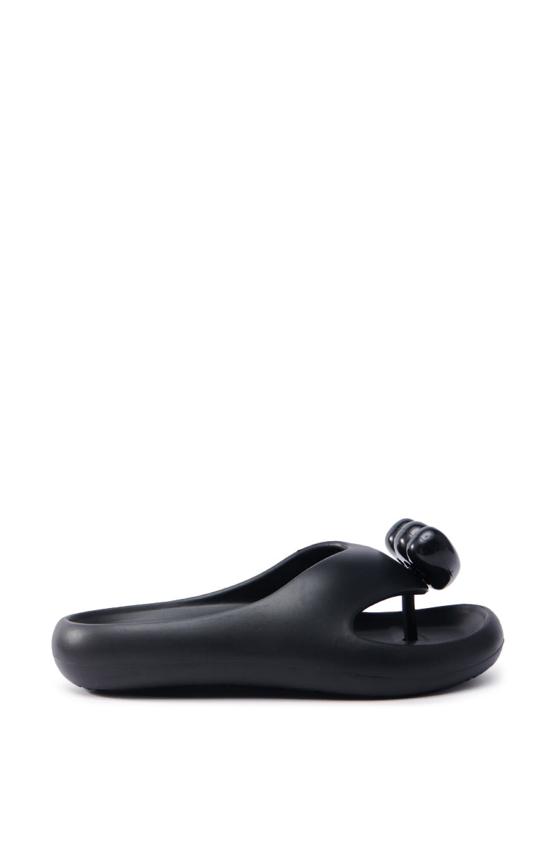 The AKIRA COMPASSION BLACK SANDAL features a unique design with an EVA upper, abstract knob-like embellishment on the front strap, and a single thong toe loop. It boasts a chunky sole and a sleek, minimalist appearance.