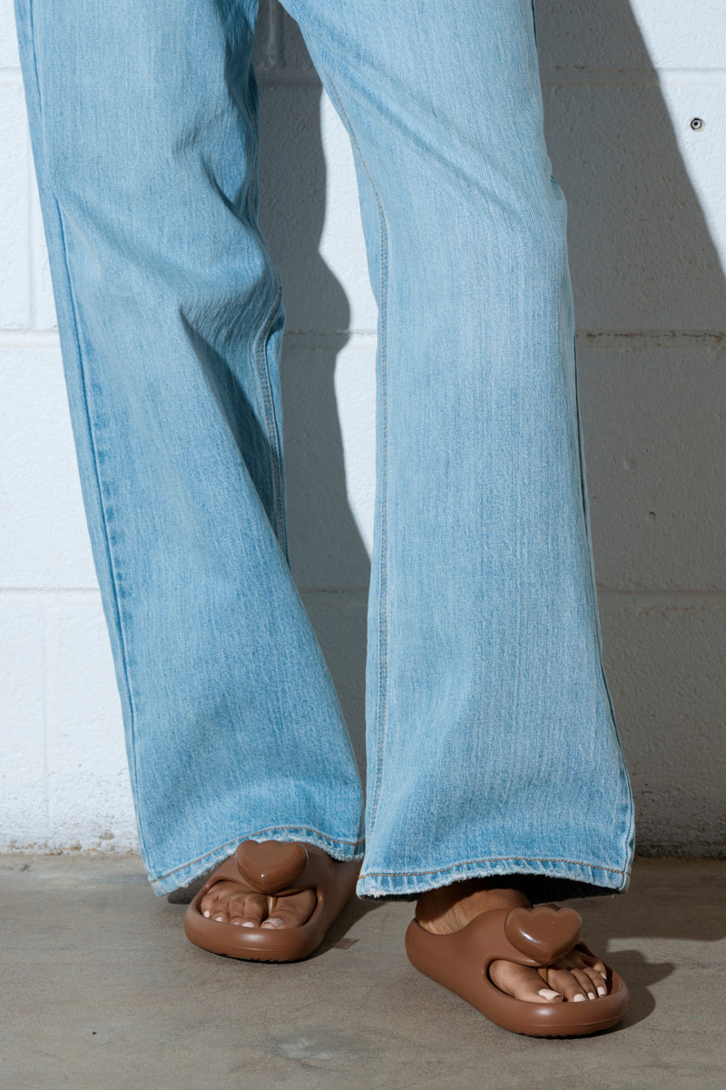 A person in light blue denim jeans is standing on a concrete floor against a light-colored wall, wearing AKIRA Compassion Brown Sandals featuring heart-shaped designs.