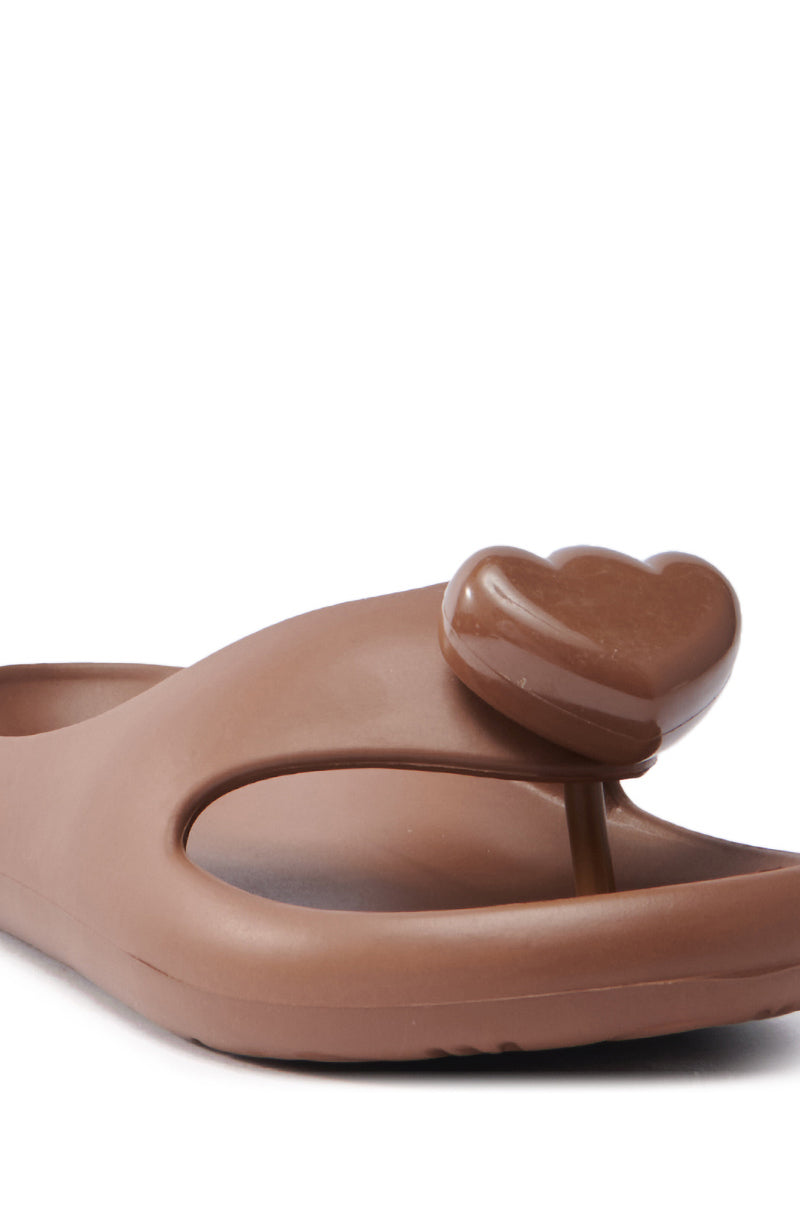 A close-up of the AKIRA COMPASSION BROWN SANDAL shows a heart-shaped detail on the almond thong toe strap. The slide-on design combines simplicity and modern style with a smooth finish.