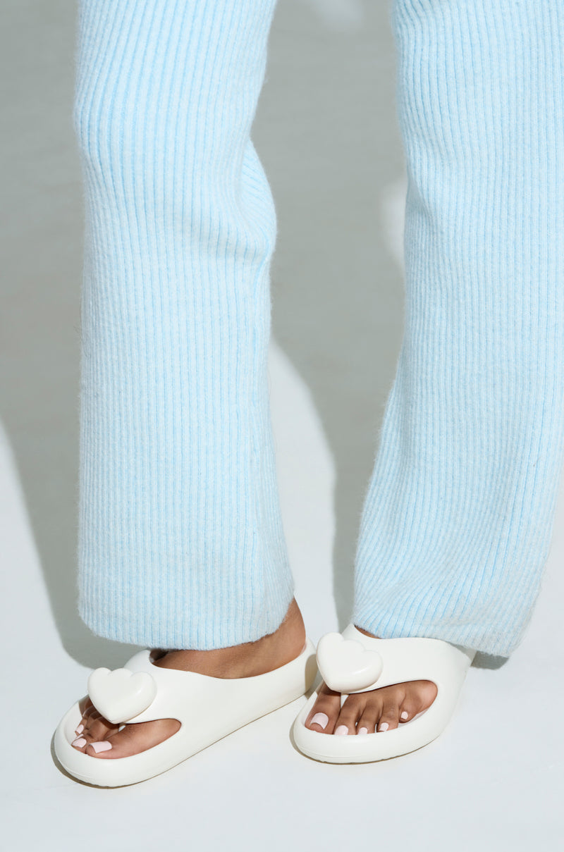 Standing on a light surface, a person enjoys the comfort underfoot while wearing light blue ribbed pants paired with AKIRA COMPASSION WHITE SANDALS, which feature heart decorations on the top.
