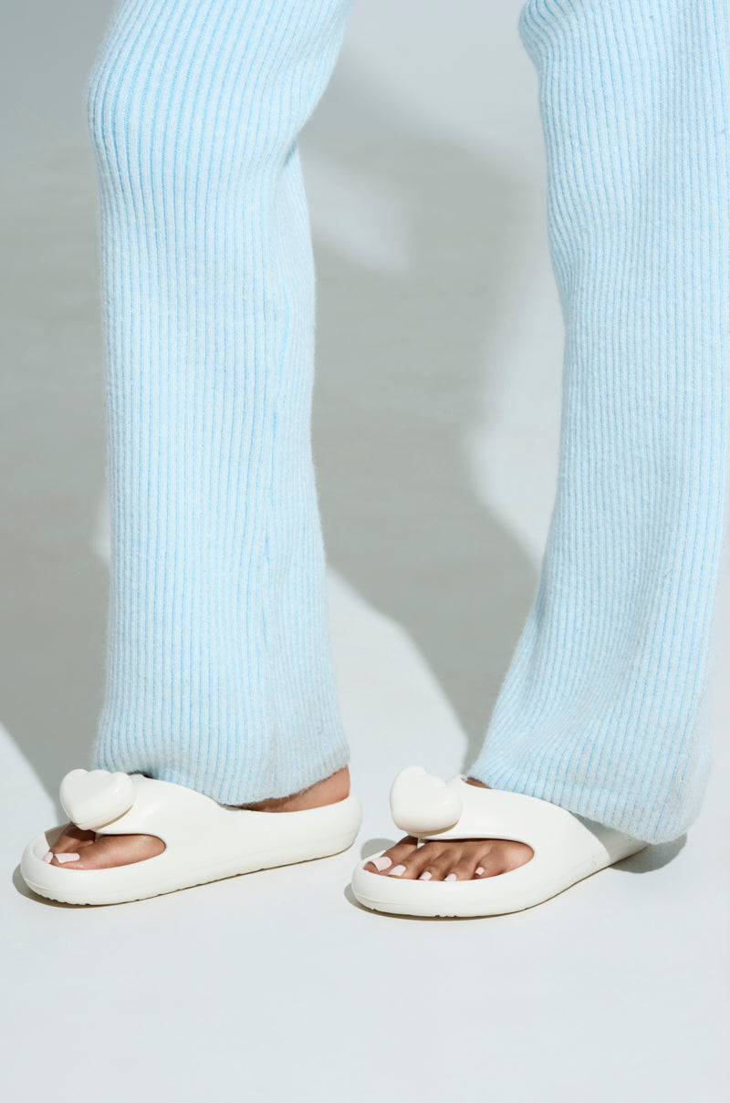A person is dressed in light blue ribbed pants and chic AKIRA COMPASSION WHITE SANDALS with heart-shaped decorations, crafted from cushy material. The sandals expose painted toenails against a plain, light-colored background.