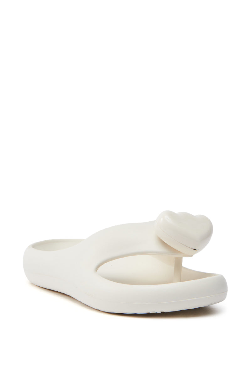 The AKIRA COMPASSION WHITE SANDAL showcases a minimalist design with a heart-shaped embellishment on the strap. Its cushy material ensures comfort, while a thick sole and almond thong toe enhance the look, all elegantly positioned on a white background.