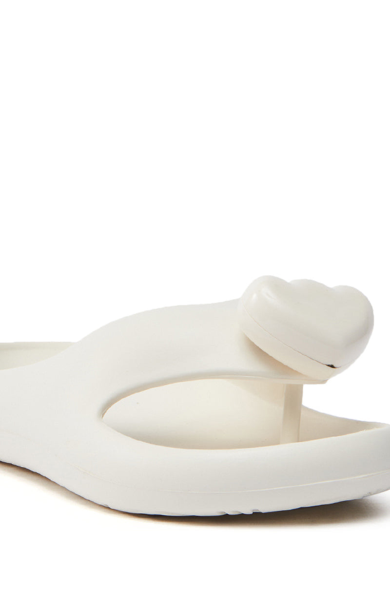 The AKIRA COMPASSION WHITE SANDAL, with an elegant heart-shaped embellishment near the almond thong toe, boasts a minimalist design of clean lines and cushy material for comfort. Photographed on a plain white background, it exemplifies chic simplicity and timelessness.