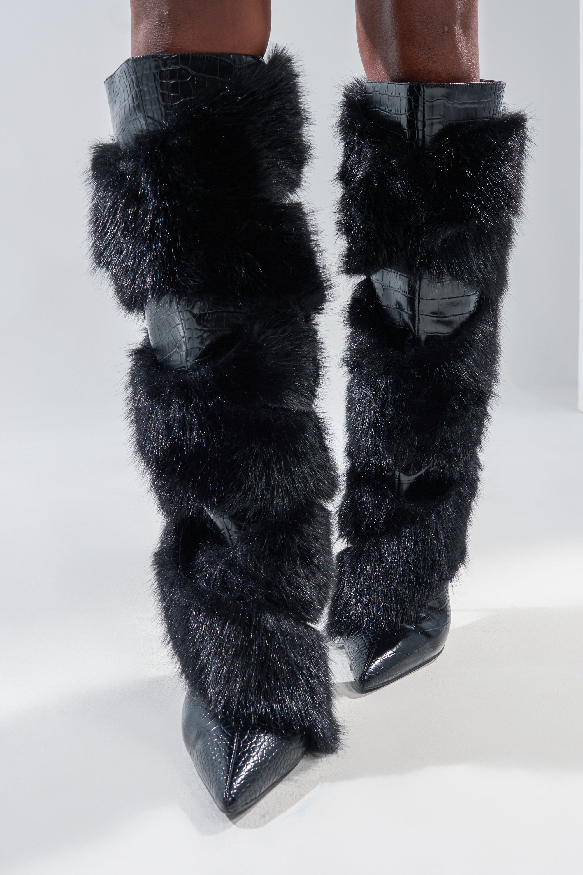 A person wearing the AZALEA WANG CONRADDE BLACK MIXED MATERIAL FUR AND SNAKE STILETTO BOOT—tall, knee-high boots with pointed toes in shiny black faux fur and leather—stands against a plain light background.