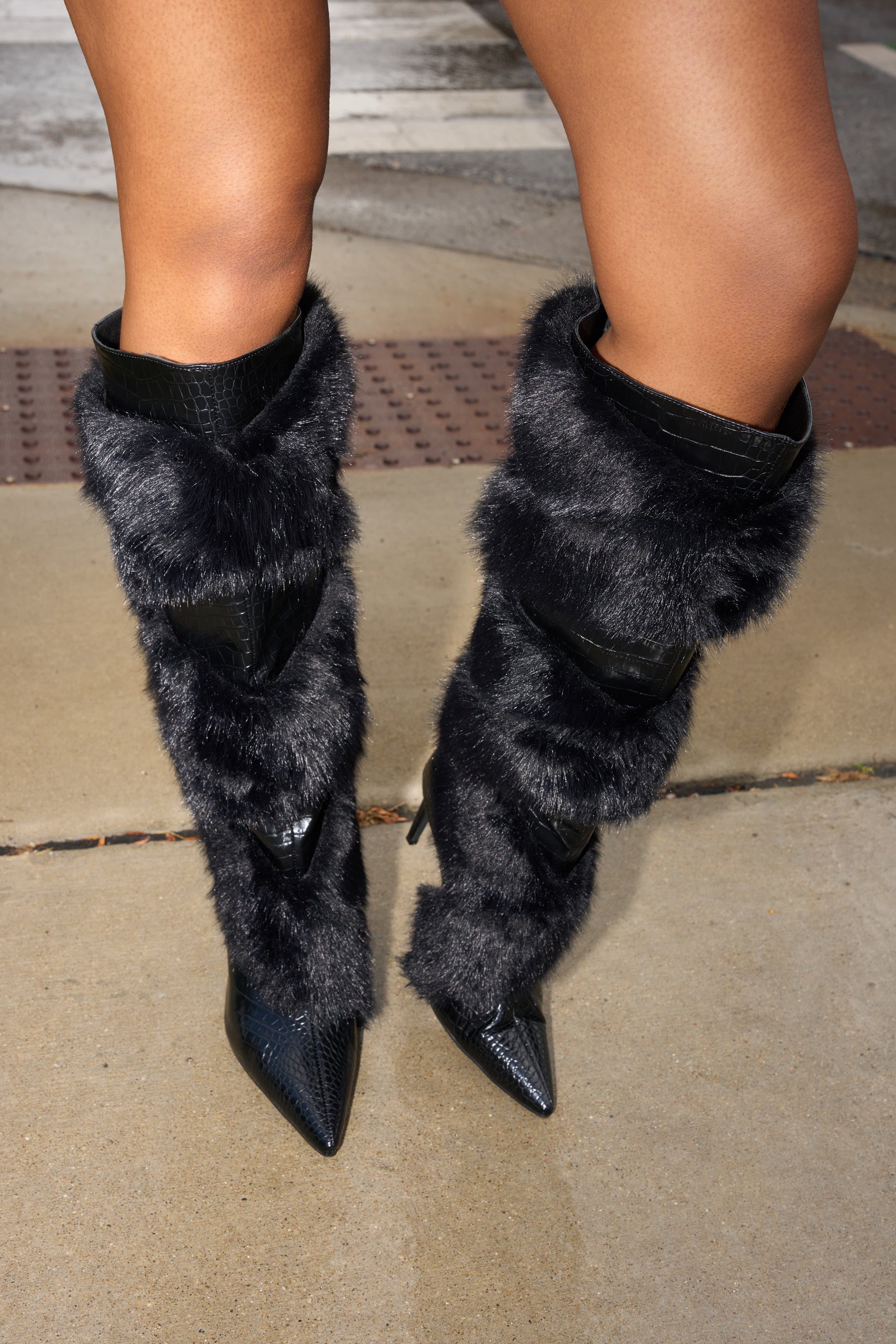 Someone wears the AZALEA WANG CONRADDE Black Mixed Material Fur and Snake Stiletto Boot, featuring pointed toes and textured faux leather details, while standing on a concrete sidewalk.