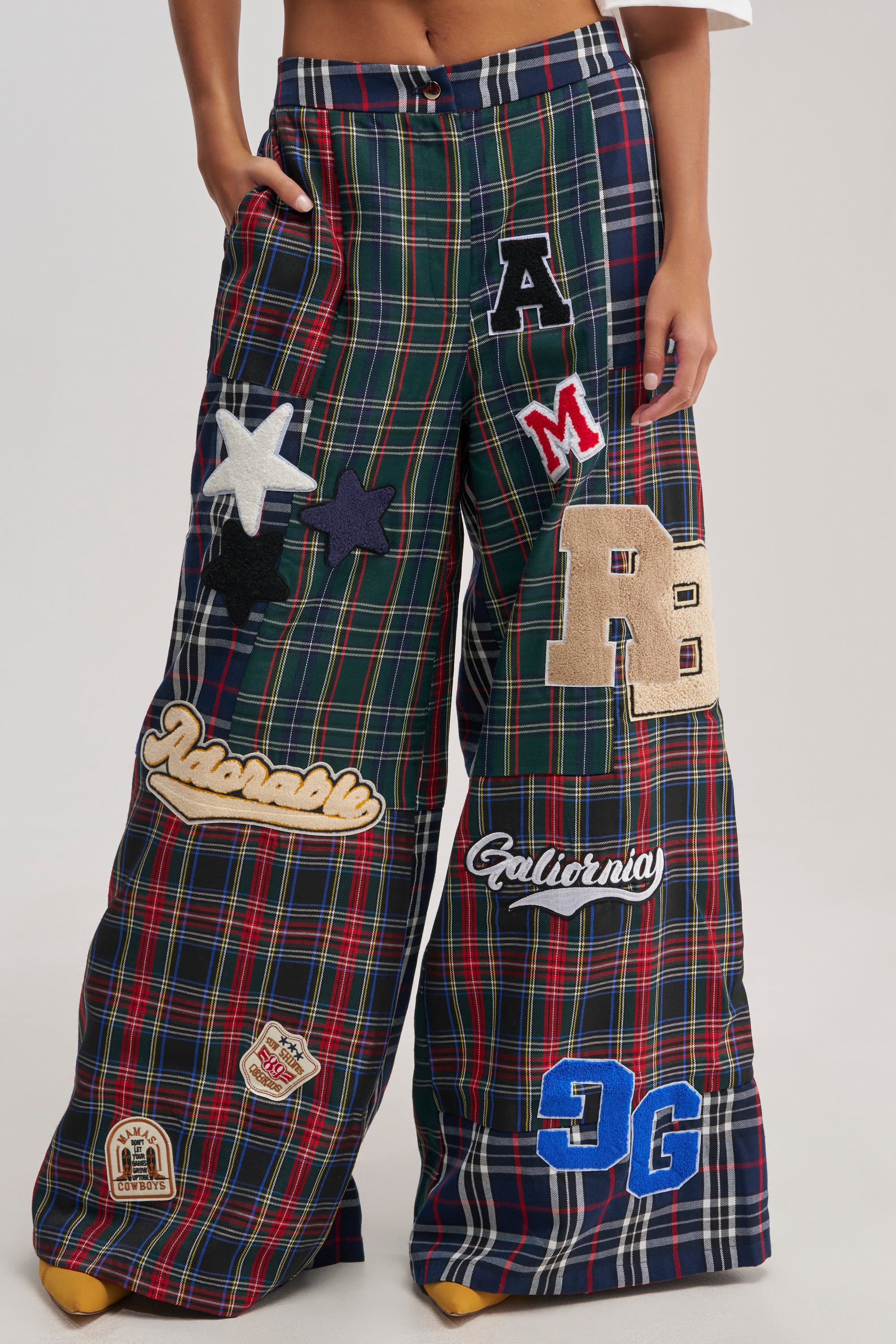 Wearing the ALL ABOUT ME PLAID TROUSER, which features high-waisted, wide-leg plaid with bold colorful patches, stars, letters, and red, green, and blue checks against a plain background.