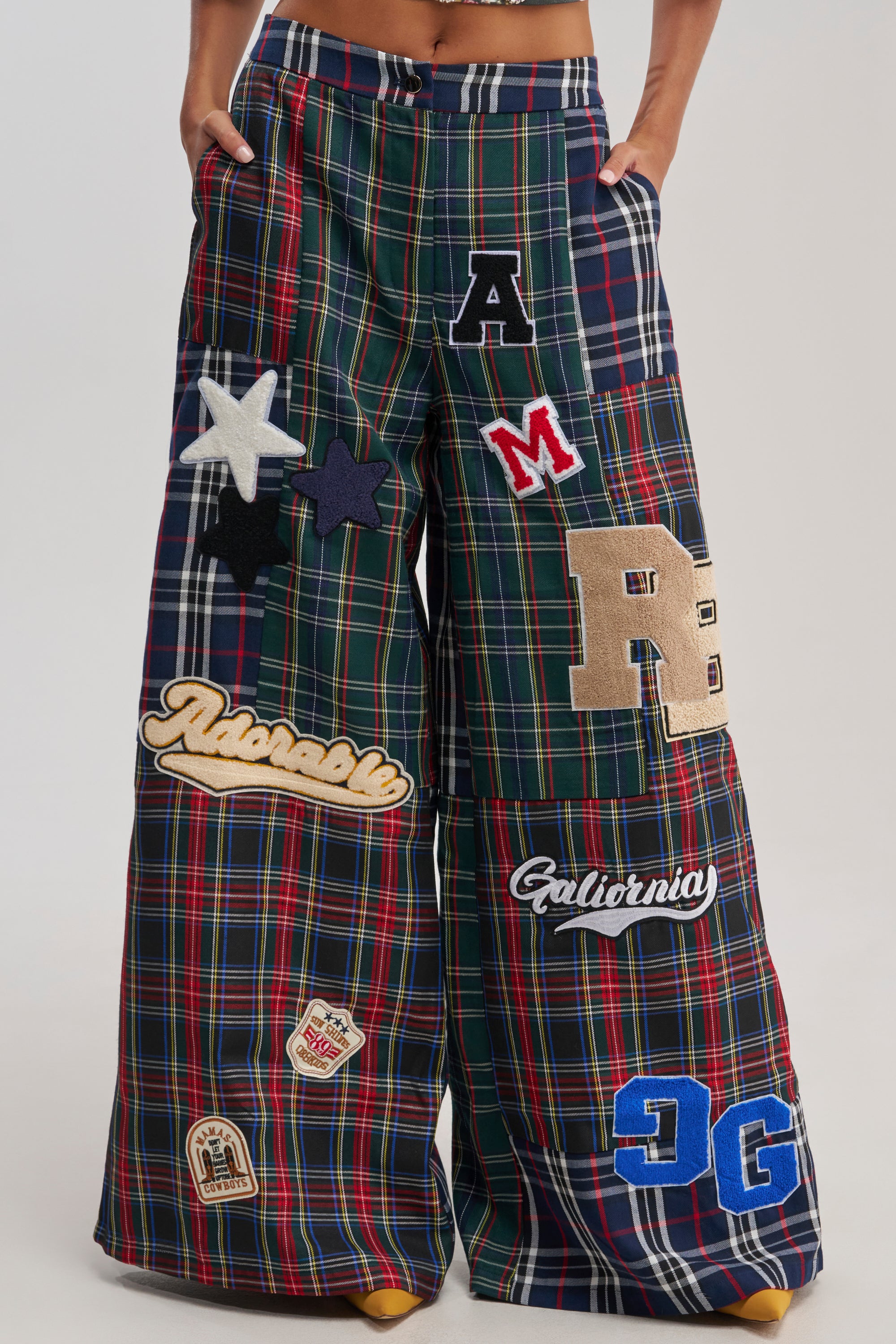 Wearing the ALL ABOUT ME PLAID TROUSER—high-waisted, wide-leg fit with large, colorful varsity patches, stars, and letters. Only the lower torso and legs are shown against a plain background.
