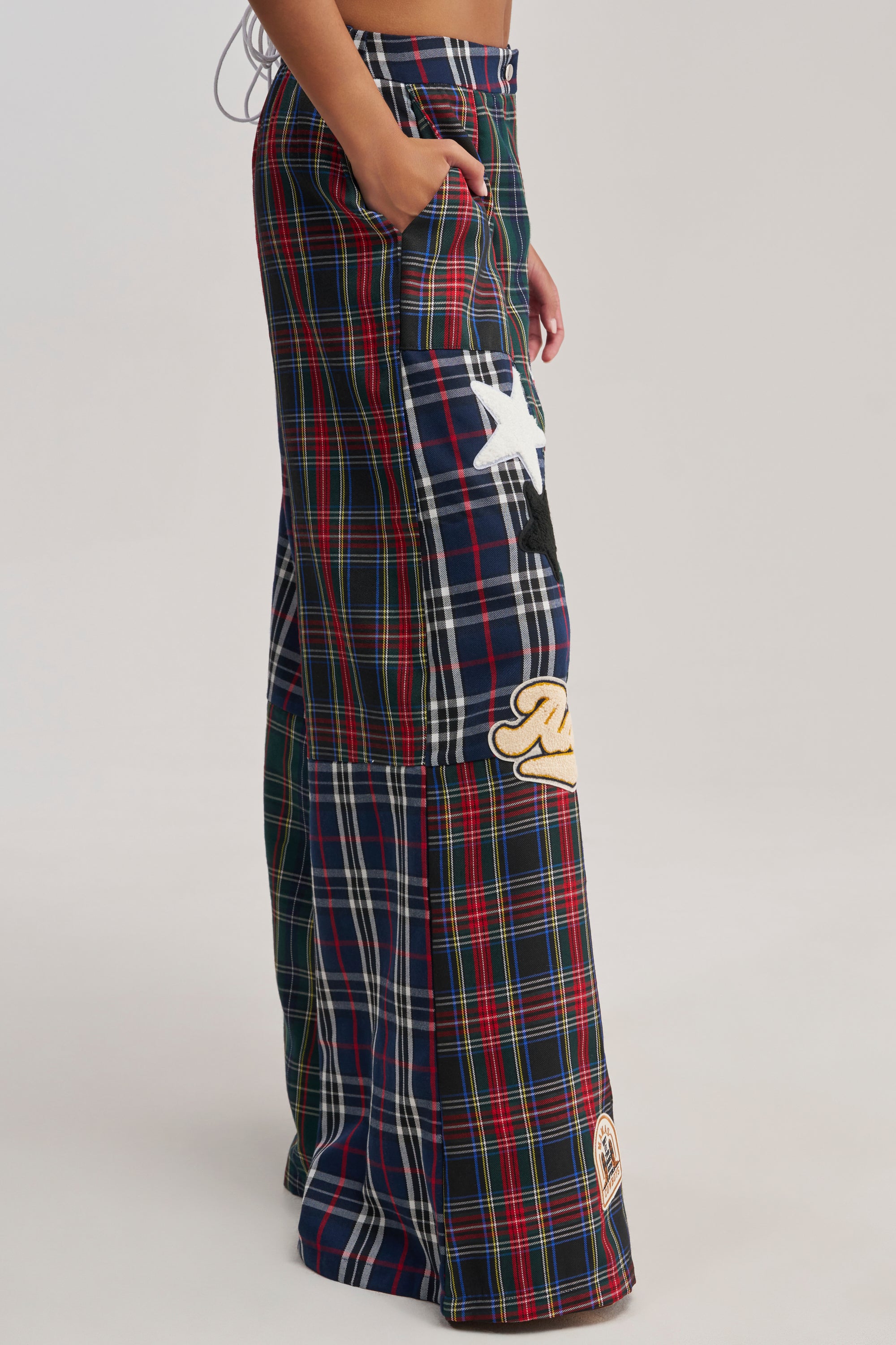 Someone stands against a plain light background wearing the ALL ABOUT ME PLAID TROUSER—high-waisted, wide-leg pants with colorful red, navy, and green plaid and varsity patch details like a white star and yellow design.