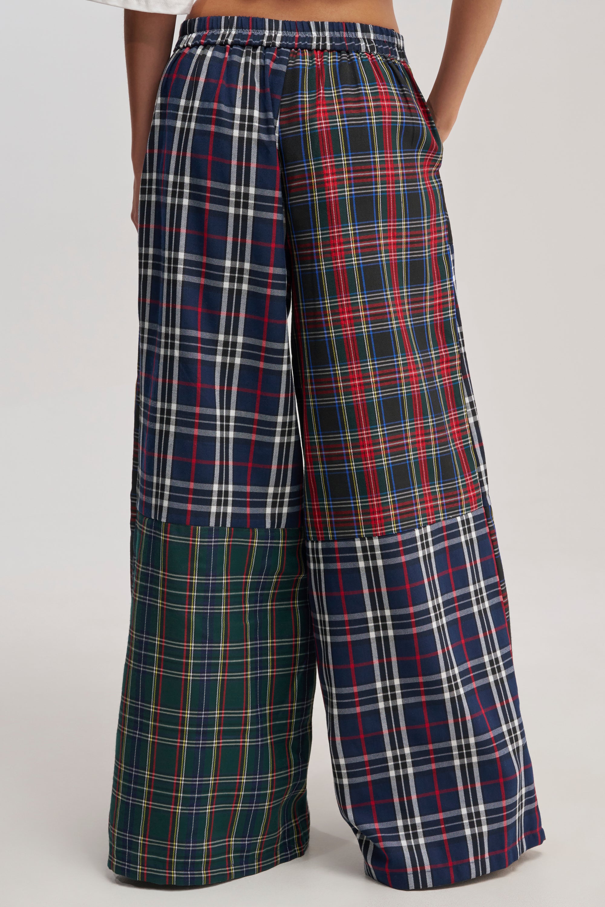 A person wears the ALL ABOUT ME PLAID TROUSER, high-waisted with a red, green, blue, and white patchwork plaid design. The cropped photo highlights the wide-leg fit from waist to feet.