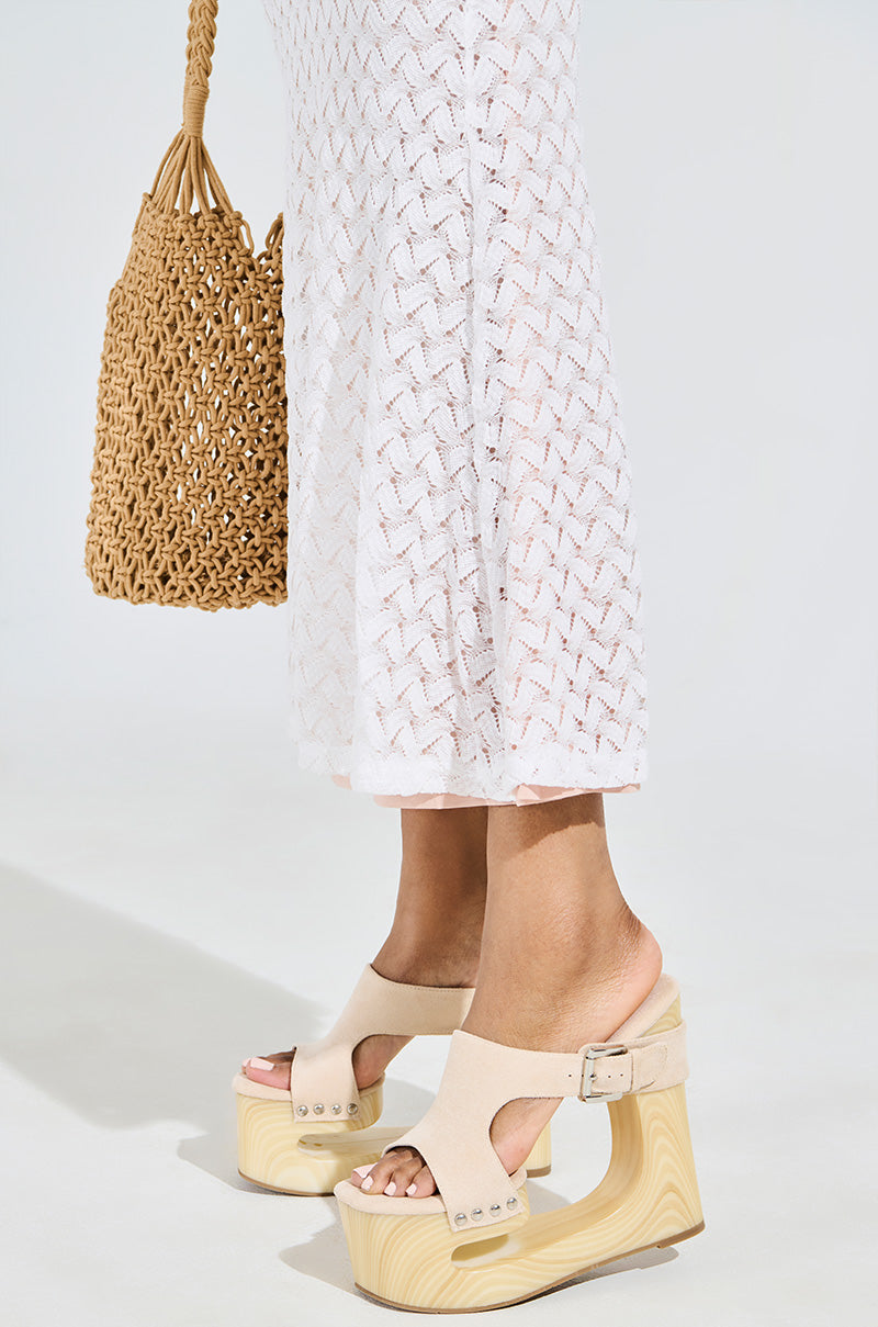 A person in a white patterned dress holds a woven bag and wears the AZALEA WANG COPSE BEIGE SANDAL, featuring faux suede with chunky wedge heels, against a plain light gray background.