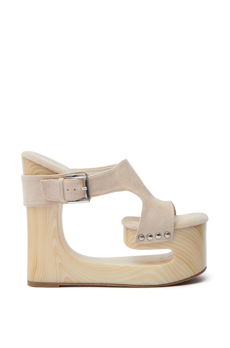 The AZALEA WANG COPSE BEIGE SANDAL showcases a stylish side view with a high-heeled light wooden wedge and beige faux suede upper. It features a chunky wide strap with a buckle and metallic studs for a modern look.