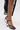 A person in a black lace dress stands on a white surface, showcasing the AZALEA WANG CORABELLA BLACK NOVELTY HEEL SANDAL featuring a faux leather upper, sculpted transparent circular heels, and ankle straps.